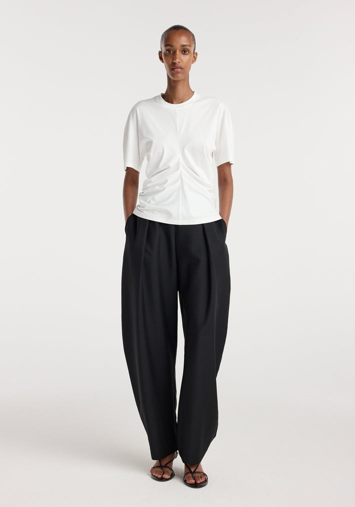 Model wearing a Draped t-shirt | off white paired with loose black trousers and black open-toe sandals, standing against a plain white background.