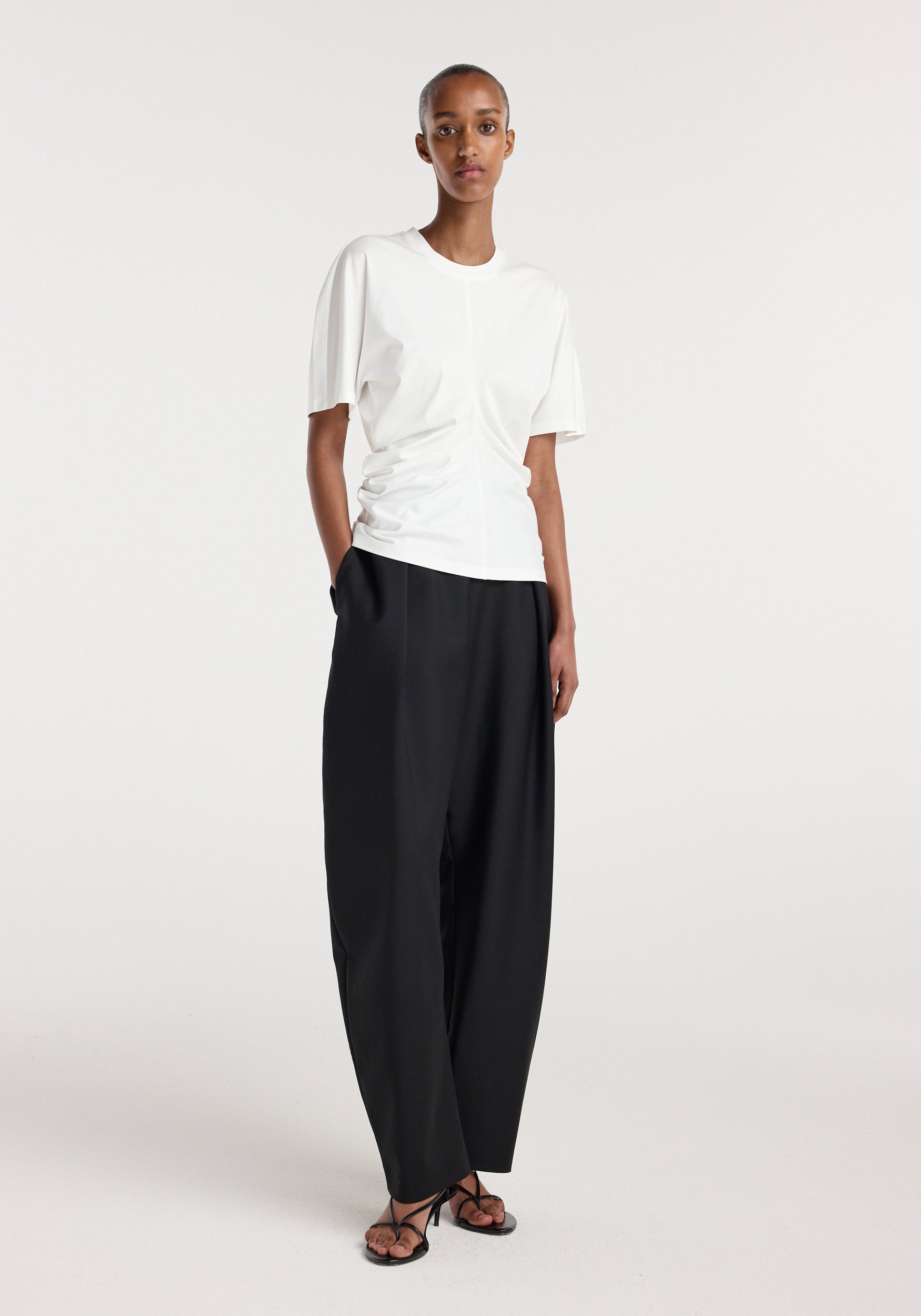 Woman wearing a fitted Draped t-shirt | off white with subtle side ruching, paired with loose black trousers and black strappy sandals, standing against a plain background.