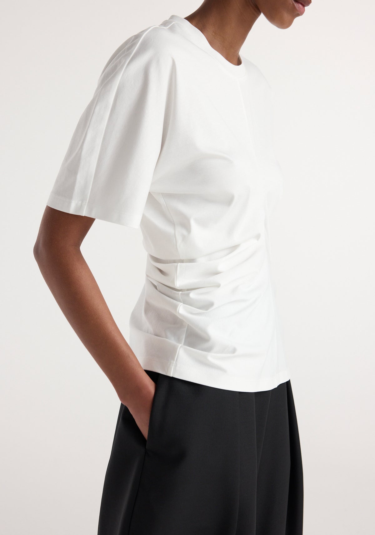 Side view of a person wearing a Draped t-shirt | off white with short sleeves and a gathered detail on the side, paired with black pants.