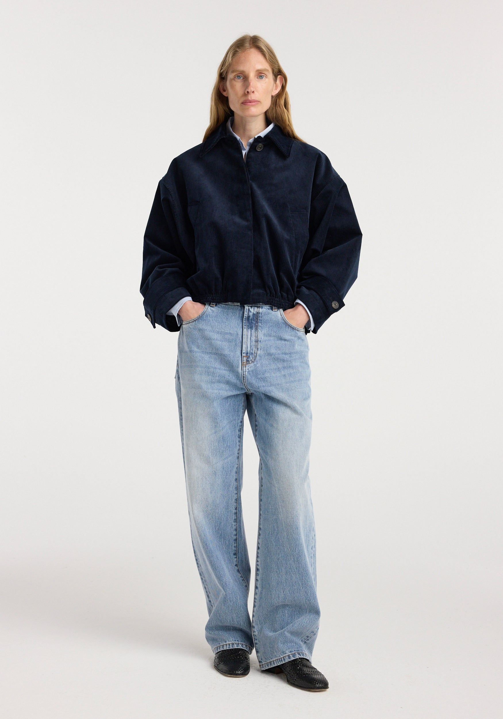 Woman wearing a dark navy corduroy blouson jacket | night with a collared shirt underneath, paired with light blue wide-leg jeans and black shoes, standing against a plain background.