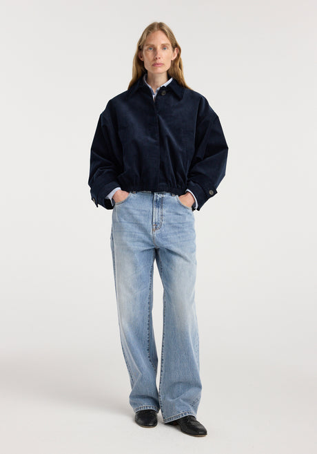 Woman wearing a dark navy corduroy blouson jacket | night with a collared shirt underneath, paired with light blue wide-leg jeans and black shoes, standing against a plain background.