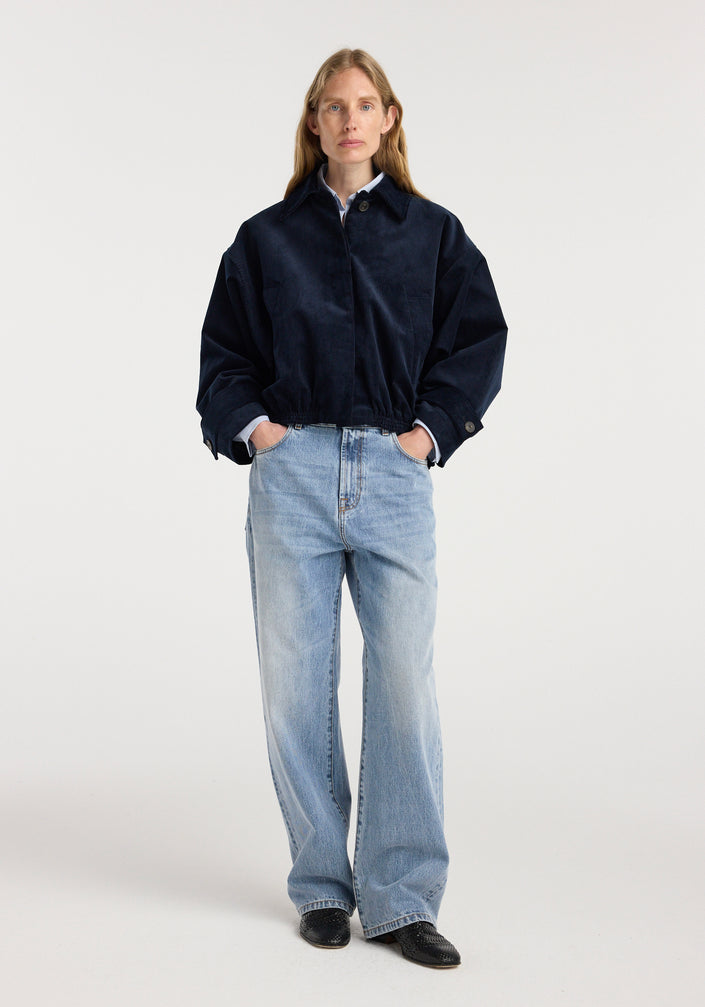Woman wearing a dark navy corduroy blouson jacket | night with a collared shirt underneath, paired with light blue wide-leg jeans and black shoes, standing against a plain background.
