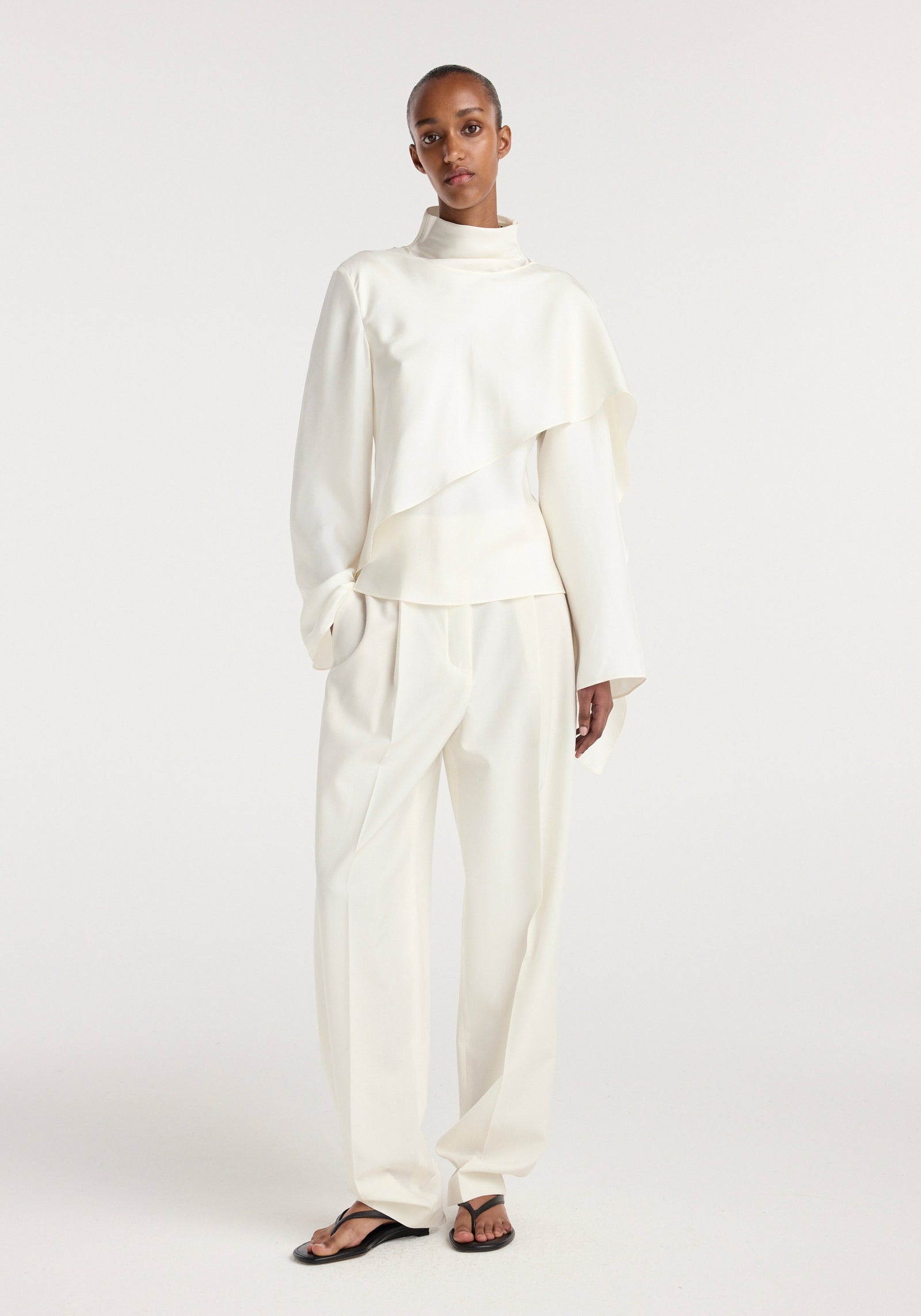 Model wearing a cream silk foulard top with long sleeves and a high neckline paired with loose cream trousers and black sandals against a plain white background.