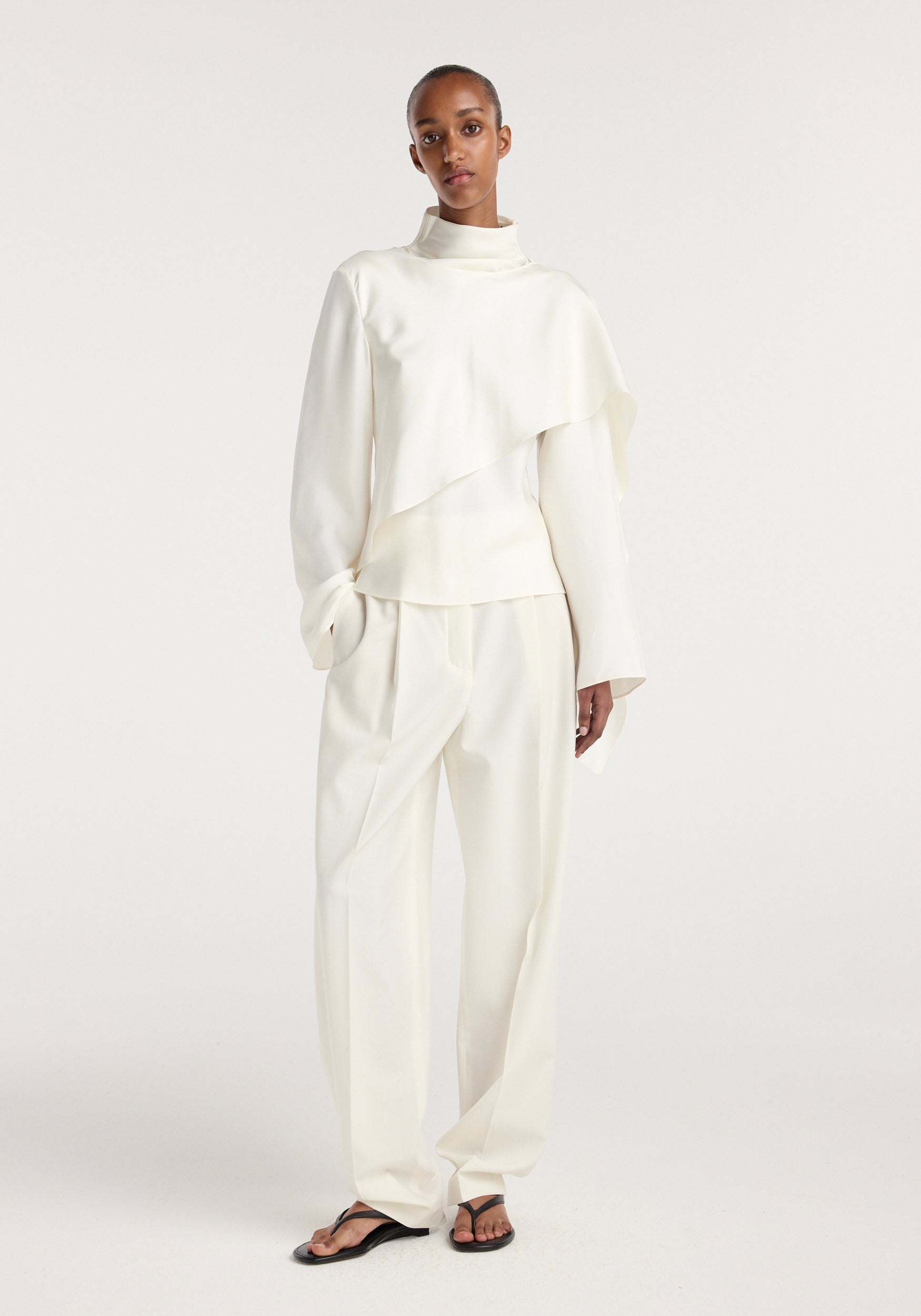 Model wearing a cream silk foulard top with long sleeves and a high neckline paired with loose cream trousers and black sandals against a plain white background.