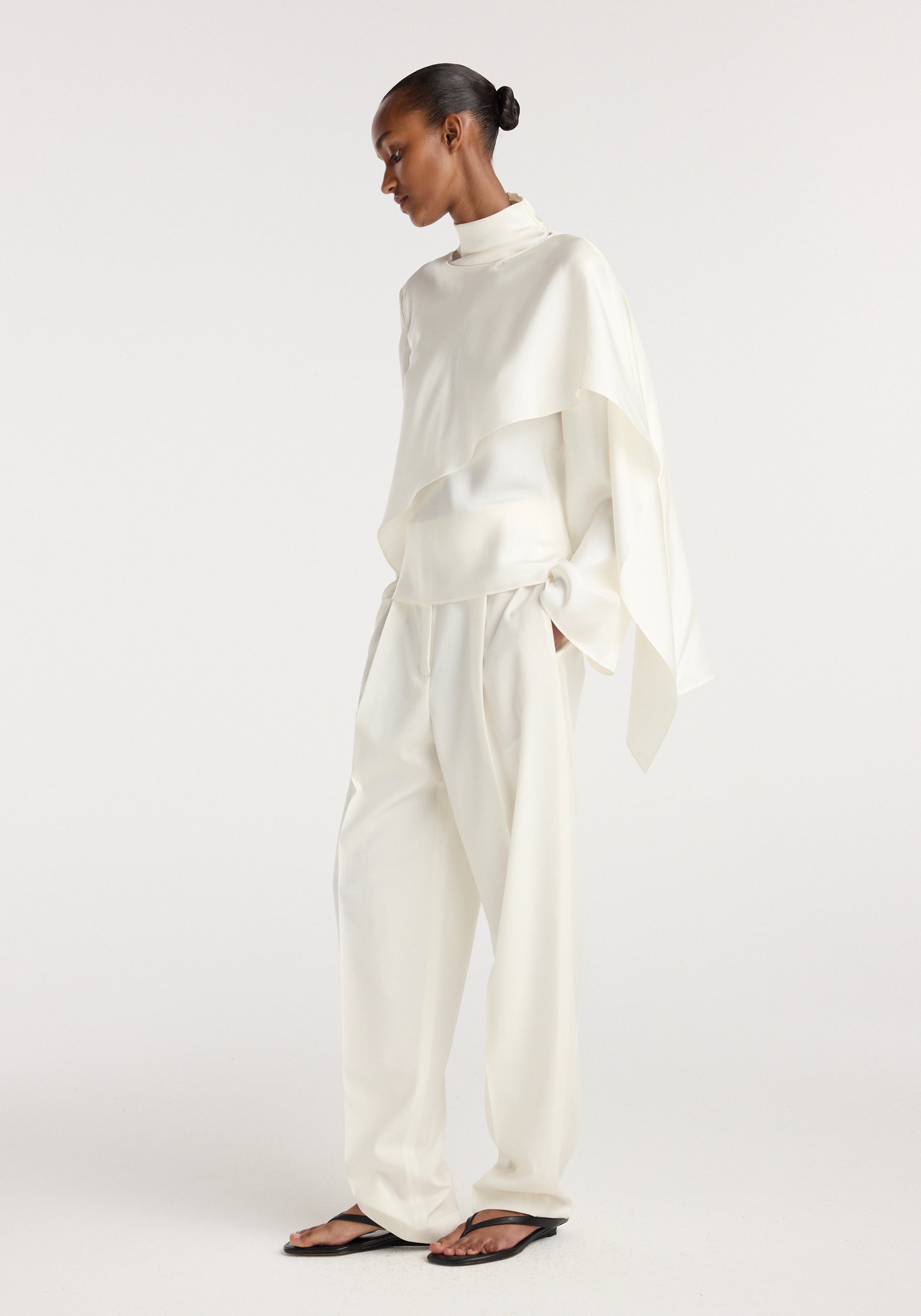 Model wearing a cream silk foulard top with a high neck and asymmetrical draped detail, paired with loose cream trousers and black sandals, standing against a plain background.