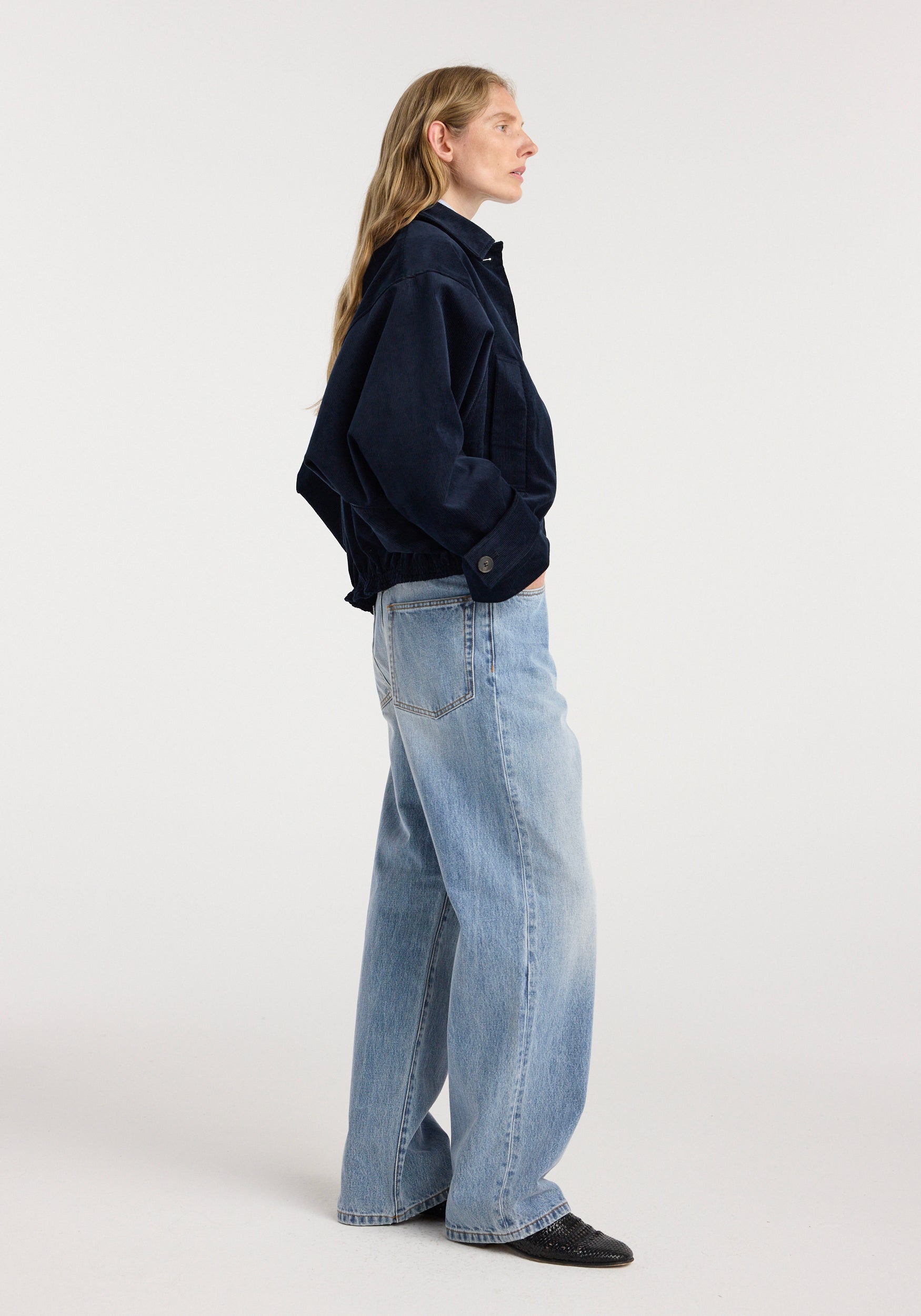 Side view of a person wearing a dark navy blue Corduroy blouson jacket | night paired with light blue wide-leg jeans and black shoes against a plain background.