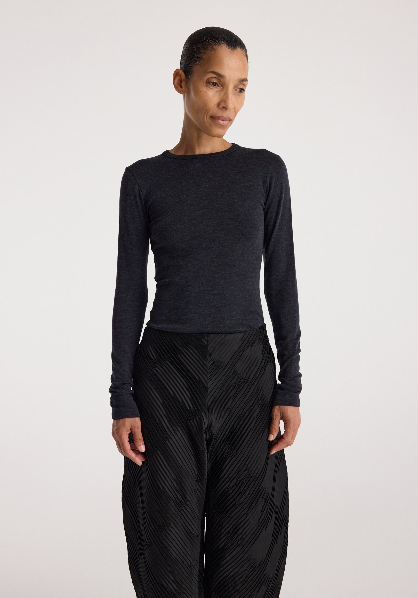 Woman wearing a fitted Fine merino long sleeve | anthracite melange top with long sleeves, paired with high-waisted black textured pants, standing against a plain background.