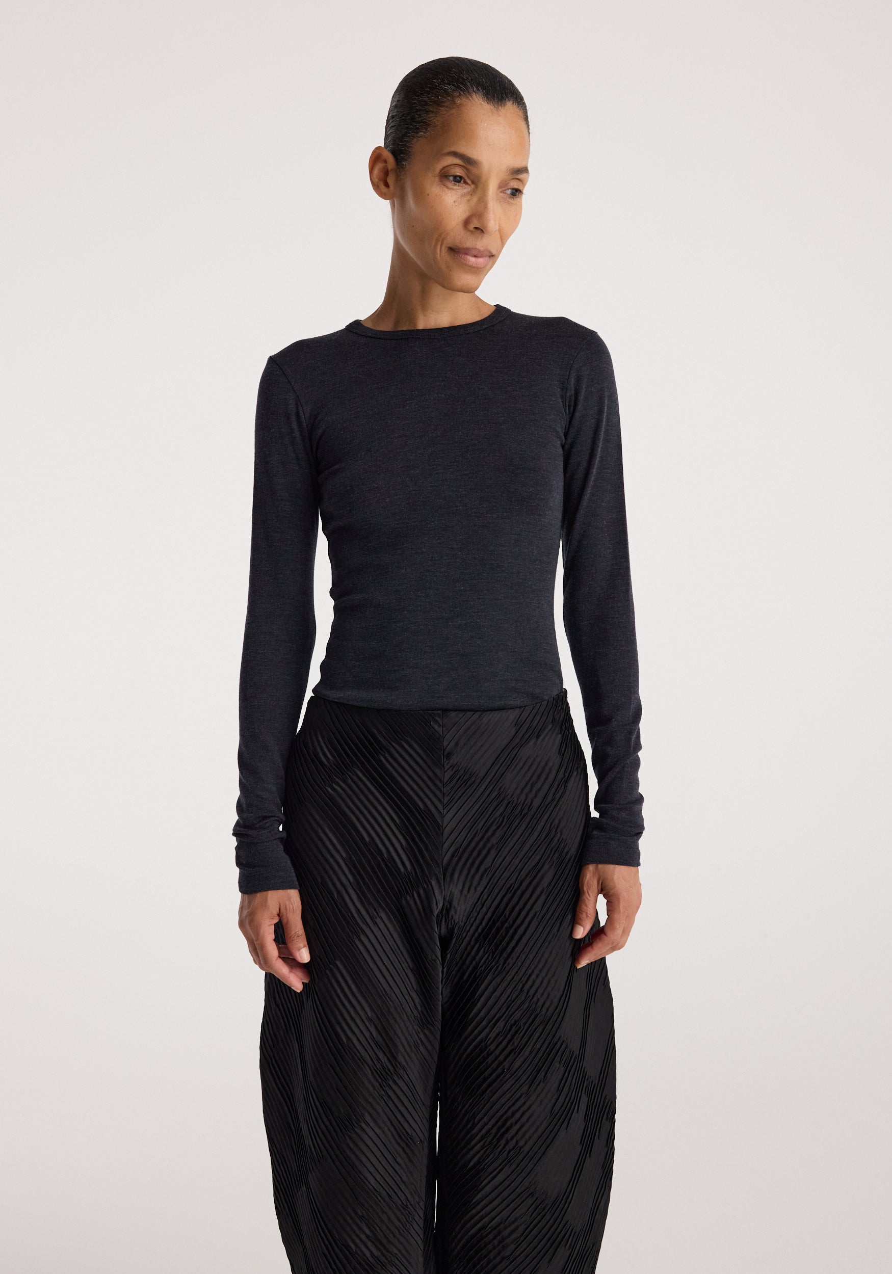 Woman wearing a fitted Fine merino long sleeve | anthracite melange top with long sleeves, paired with high-waisted black textured pants, standing against a plain background.