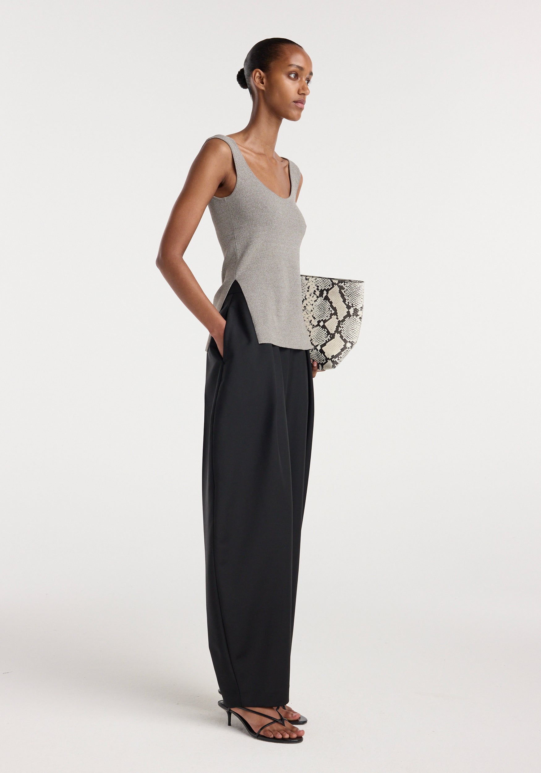 Model wearing a taupe melange long knitted bustier top paired with high-waisted black wide-leg trousers and black strappy heels, holding a snakeskin print clutch bag.