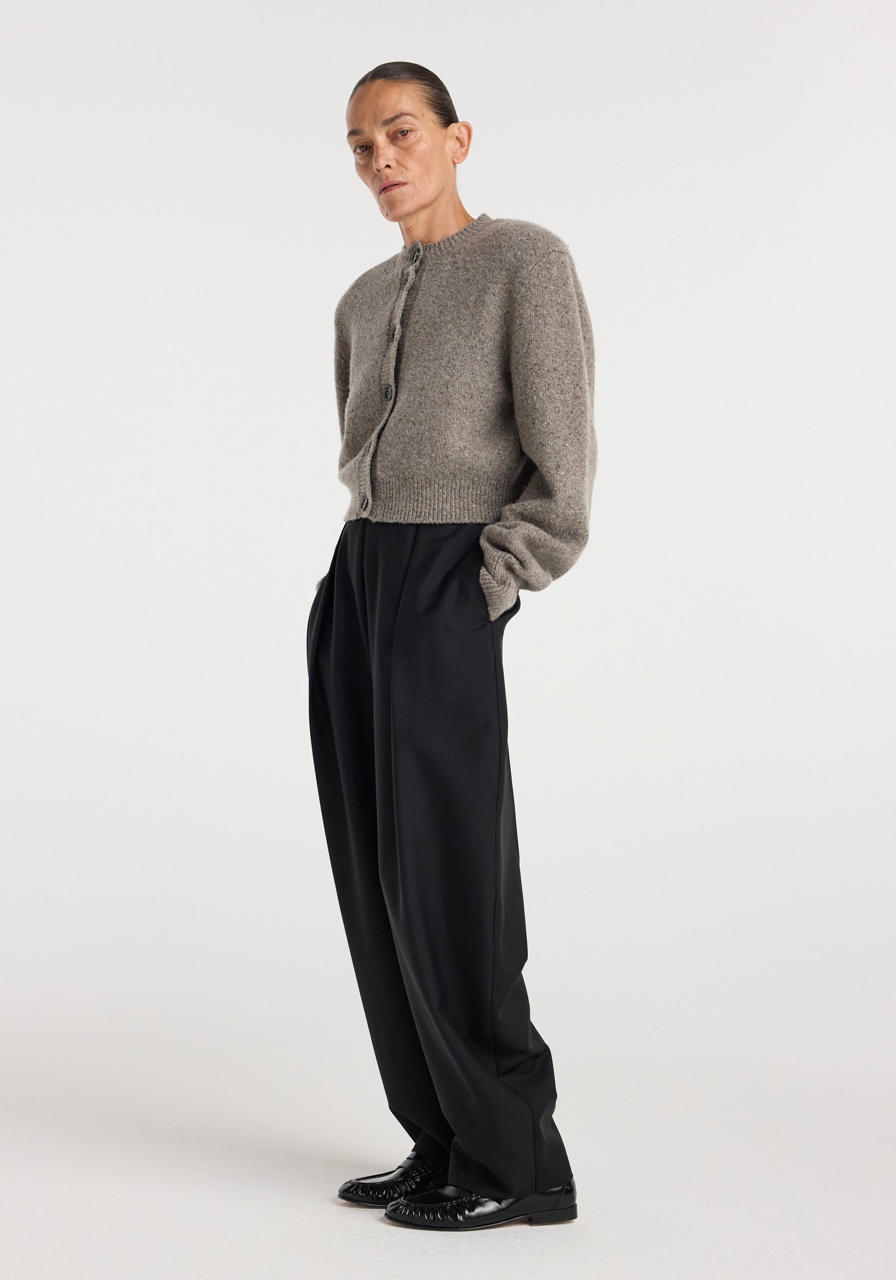 Woman wearing a cozy, cropped Statement mouliné cardigan | dark taupe melange with long sleeves and button closure, paired with wide-leg black pants and black shoes against a plain background.