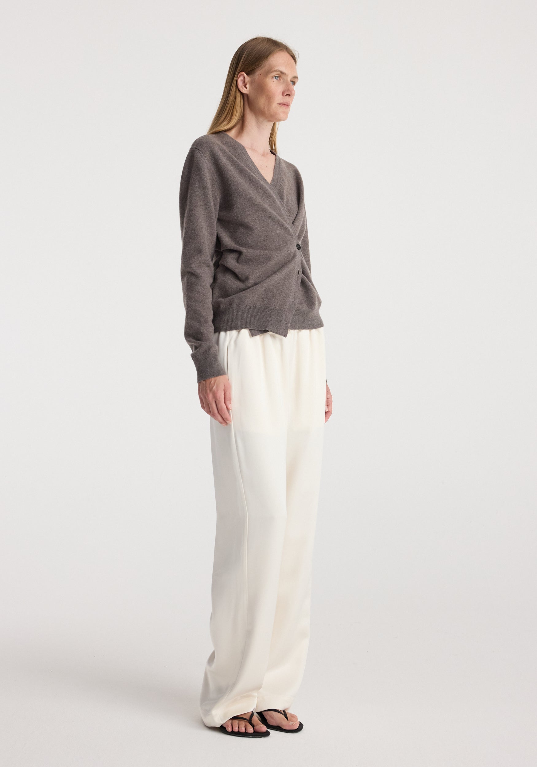 Woman wearing a twisted ballet cardigan | mud in a soft brown shade with a wrap-style front and button detail, paired with loose, white wide-legged pants and black sandals.