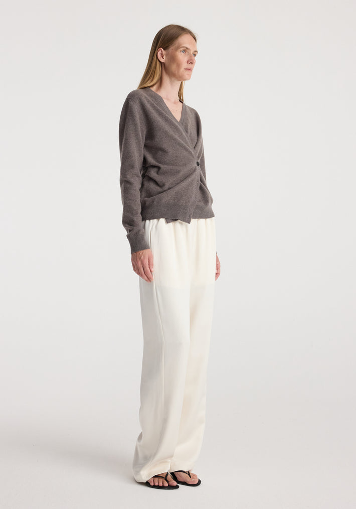 Woman wearing a twisted ballet cardigan | mud in a soft brown shade with a wrap-style front and button detail, paired with loose, white wide-legged pants and black sandals.