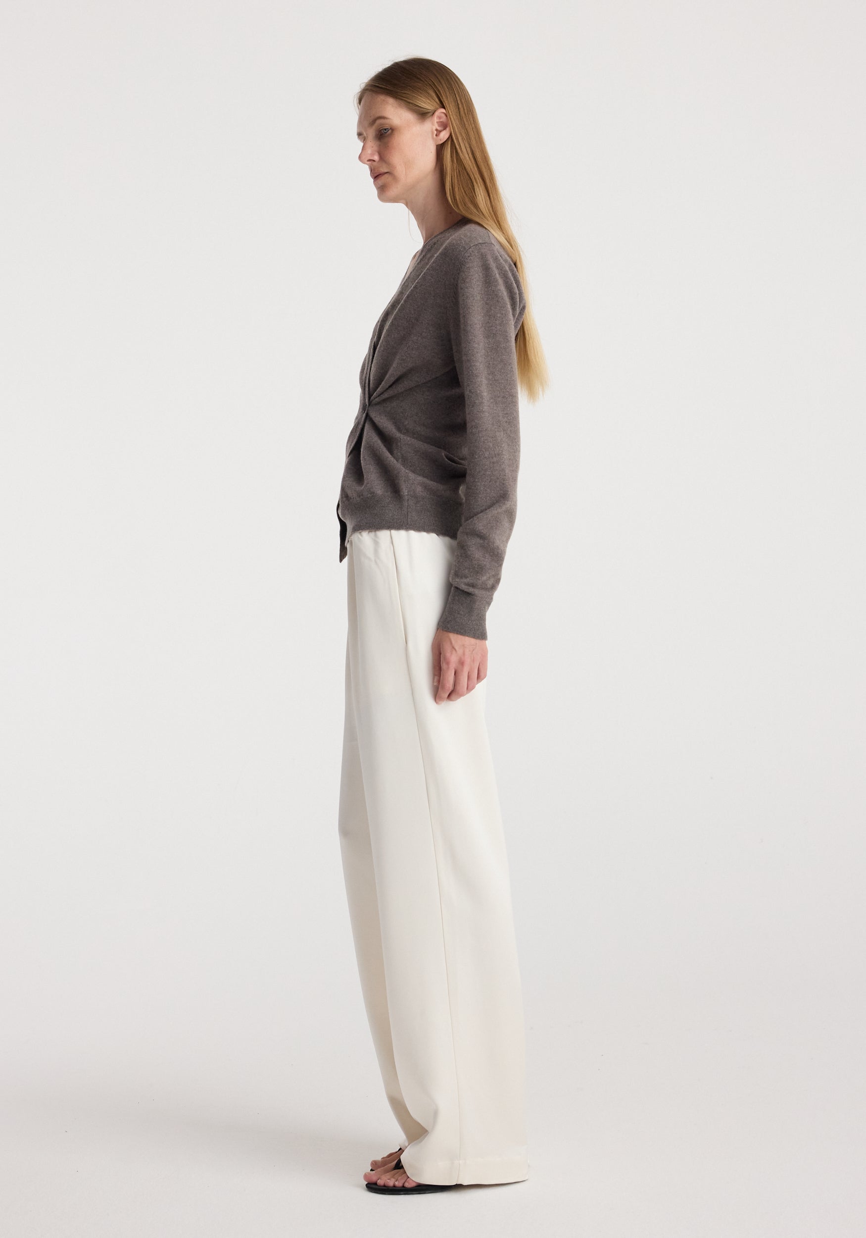Side profile of a woman wearing a twisted ballet cardigan | mud in a muted brown shade paired with loose white trousers and black sandals against a plain background.