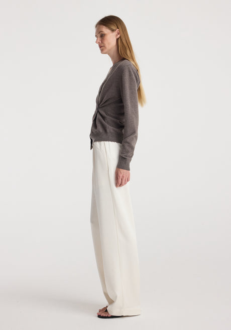 Woman wearing a twisted ballet cardigan | mud in a soft brown shade with a wrap-style front and button detail, paired with loose, white wide-legged pants and black sandals.