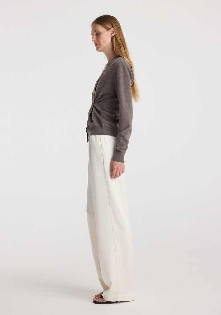 Side profile of a woman wearing a twisted ballet cardigan | mud in a muted brown shade paired with loose white trousers and black sandals against a plain background.