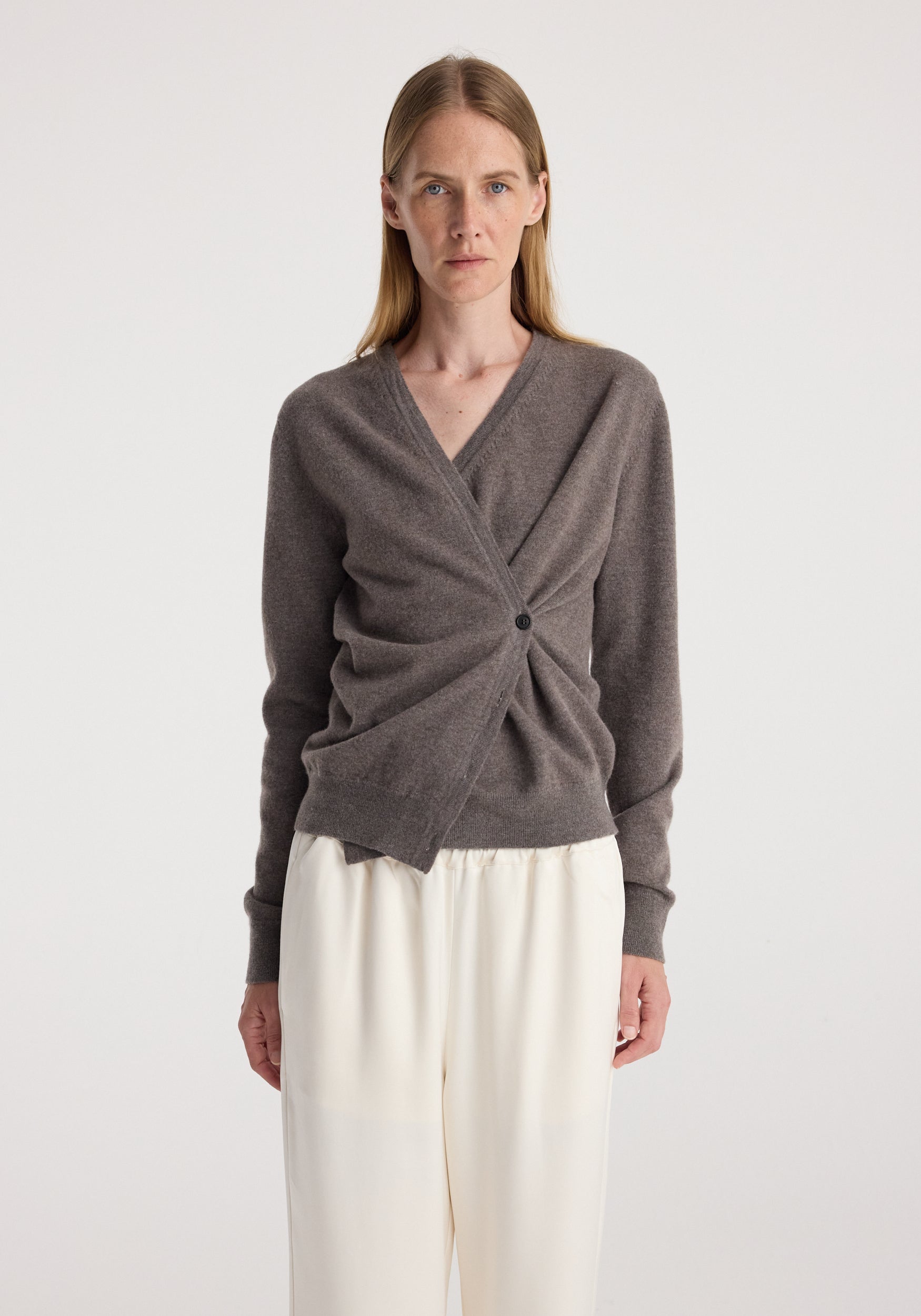 Woman wearing a twisted design wrap cardigan in a muted brown shade paired with loose white pants, styled simply against a plain background, showcasing the Twisted ballet cardigan | mud.