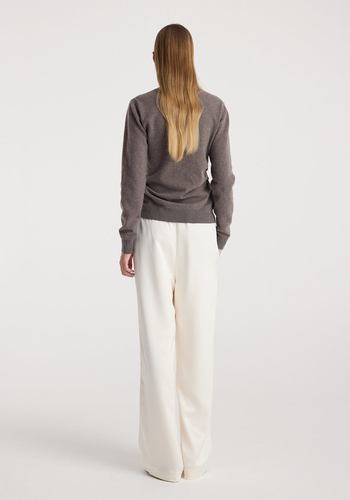 Woman seen from behind wearing a twisted ballet cardigan | mud in a muted brown shade paired with loose white pants against a plain white background.