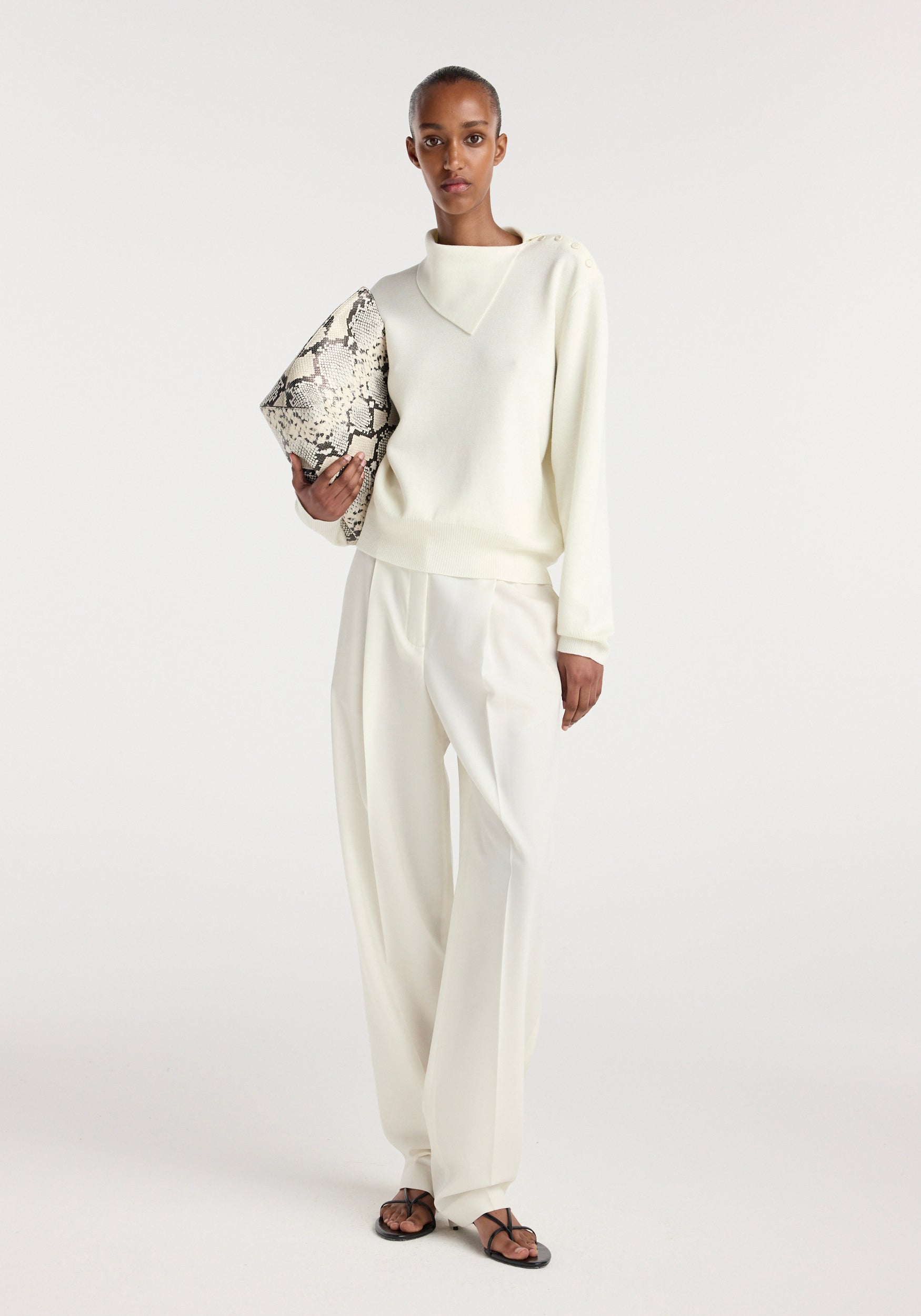 Model wearing an off white uneven collar polo paired with matching wide-leg trousers, holding a snakeskin-patterned clutch bag and wearing black strappy sandals.
