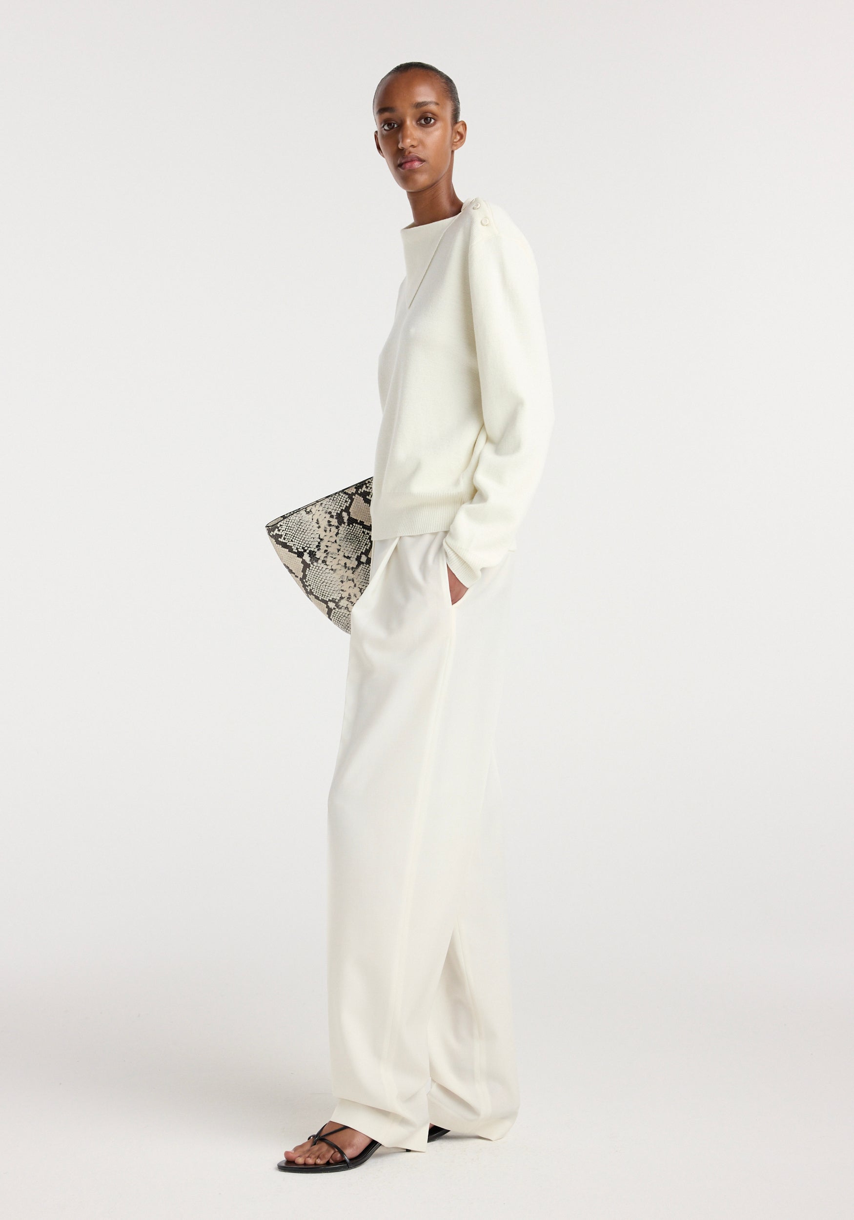 Model wearing an off white uneven collar polo paired with loose off white trousers, black sandals, and carrying a snakeskin patterned clutch against a plain background.