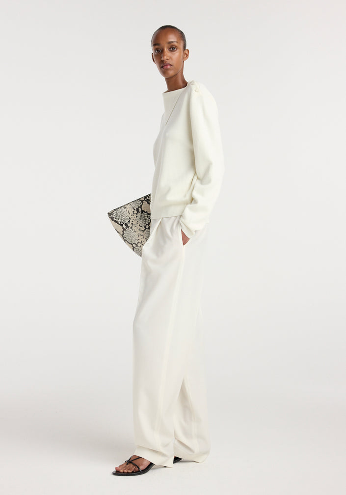 Model wearing an off white uneven collar polo paired with matching wide-leg trousers, holding a snakeskin-patterned clutch bag and wearing black strappy sandals.