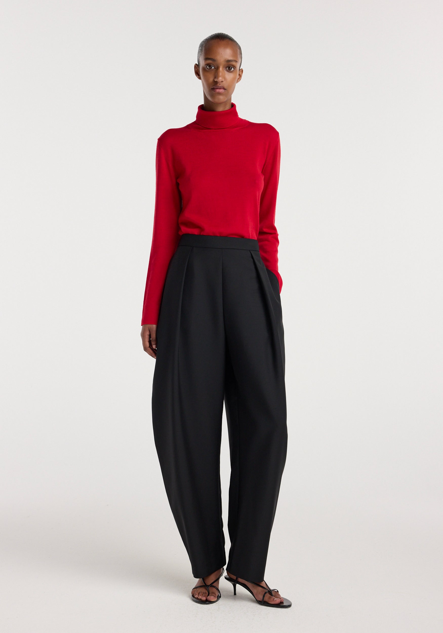 Model wearing Puck - Sculptural tailored trousers | black styled with a red long-sleeve turtleneck and black heeled sandals against a plain light background.
