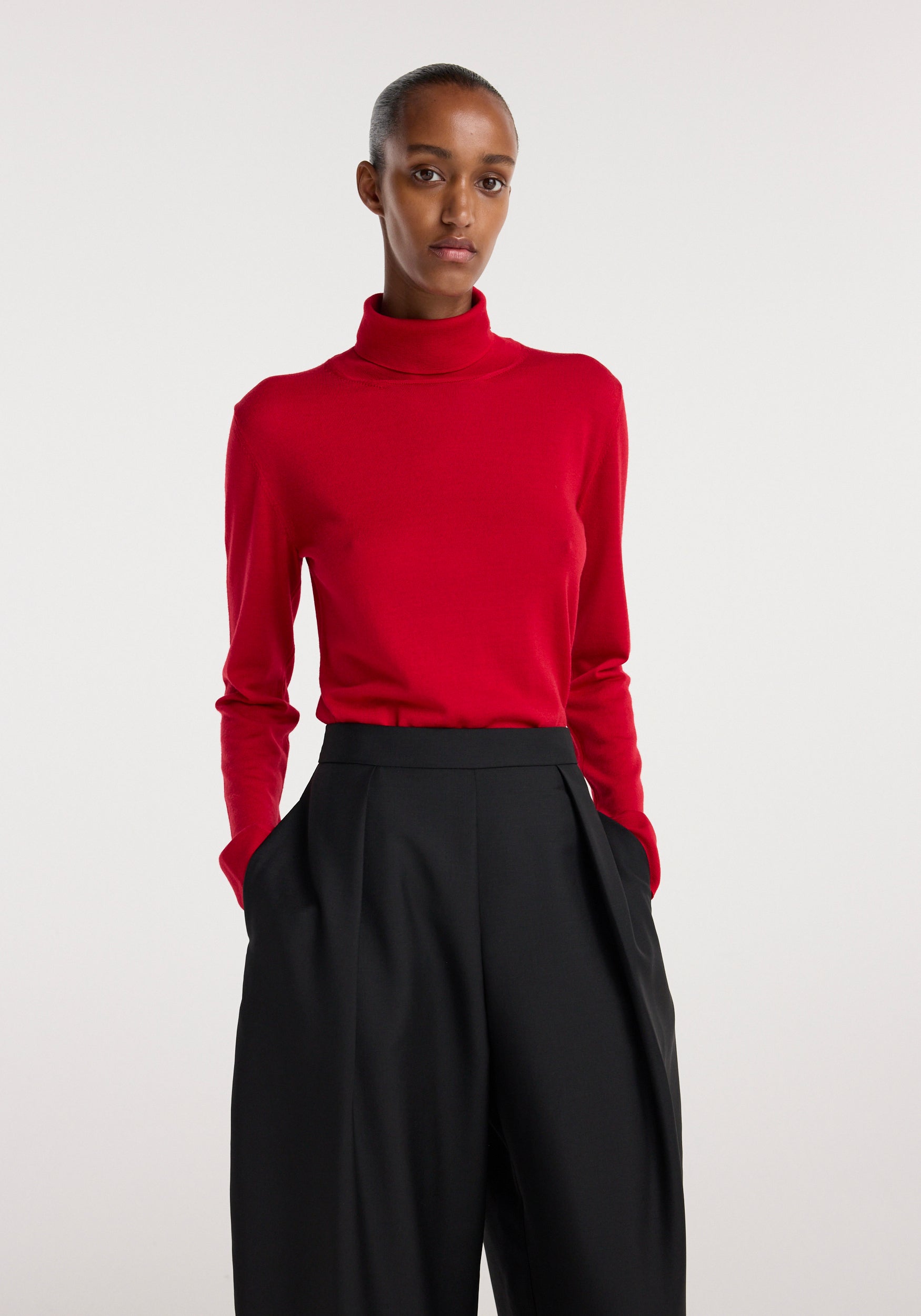 Model wearing a red fine-knit wool cashmere turtleneck with long sleeves tucked into high-waisted black wide-leg trousers against a plain background.