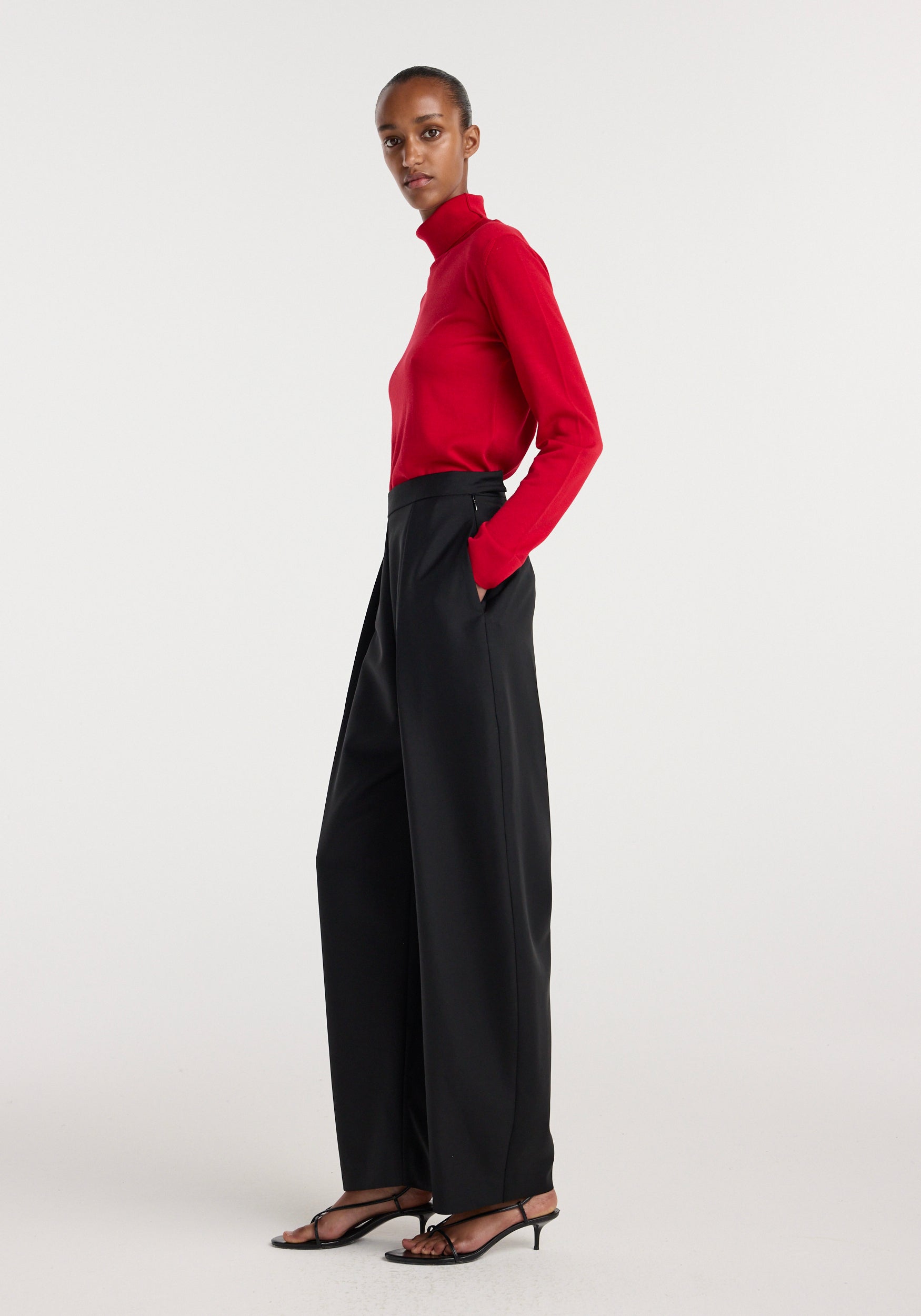 Woman wearing black Puck - Sculptural tailored trousers | black with a high waist and pleated front, paired with a red long-sleeve turtleneck and black strappy heels, standing in profile.