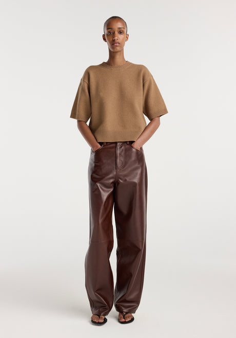 A woman wearing a camel-colored compact knitted top paired with high-waisted, wide-leg dark brown leather pants and black sandals, standing against a plain background.