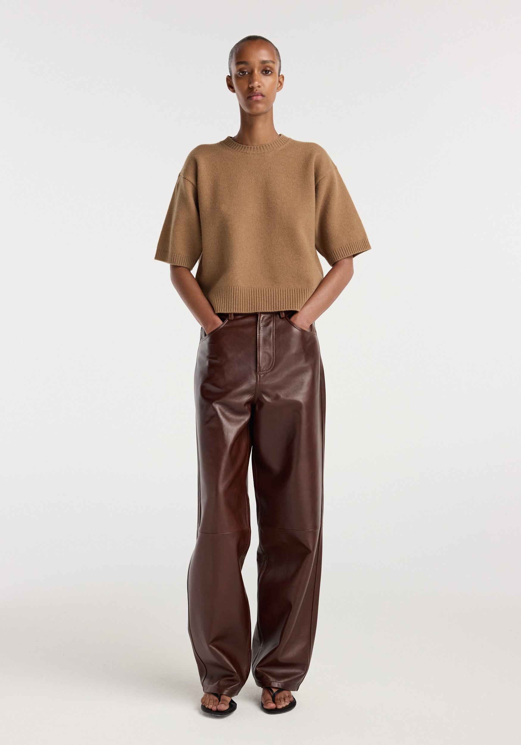 A model wearing Bruun - Signature baggy leather trousers | tobacco paired with a short-sleeve camel-colored knit sweater and black open-toe sandals, standing against a plain white background.
