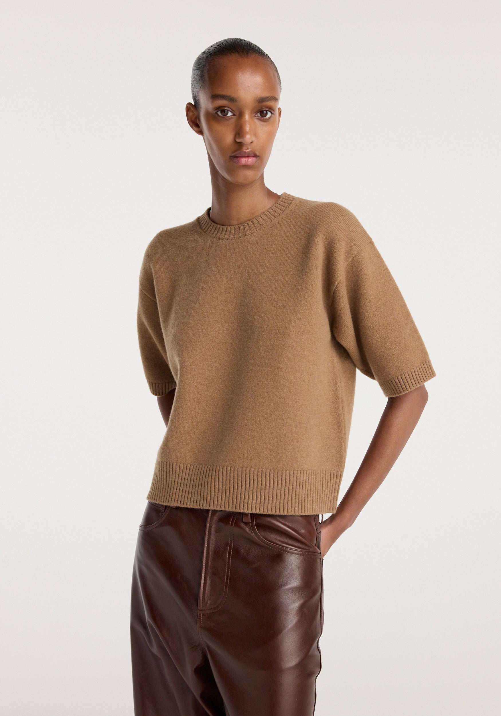 Woman wearing a camel-colored Compact knitted top | camel with short sleeves and a round neckline, paired with dark brown leather pants, standing against a plain light background.