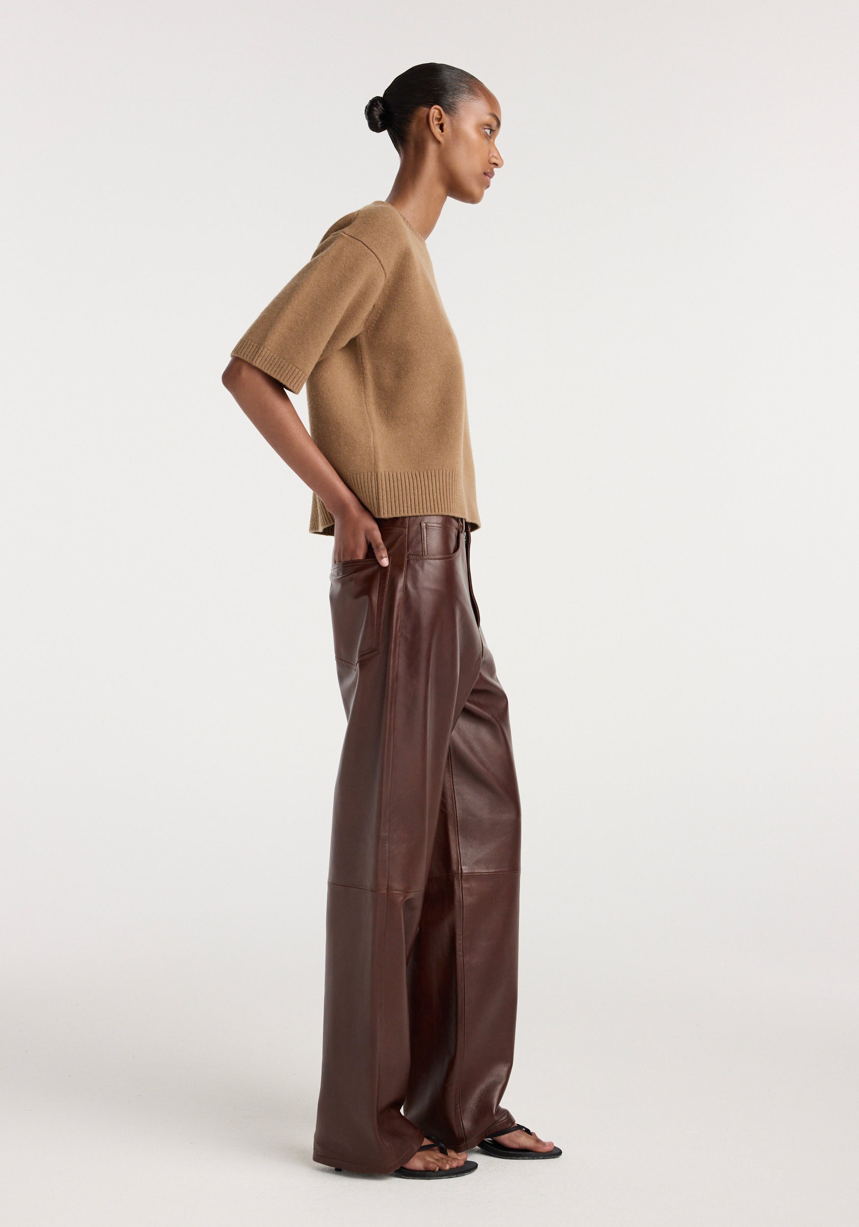 Side view of a woman wearing Bruun - Signature baggy leather trousers | tobacco, paired with a beige short-sleeve sweater and black sandals against a plain background.