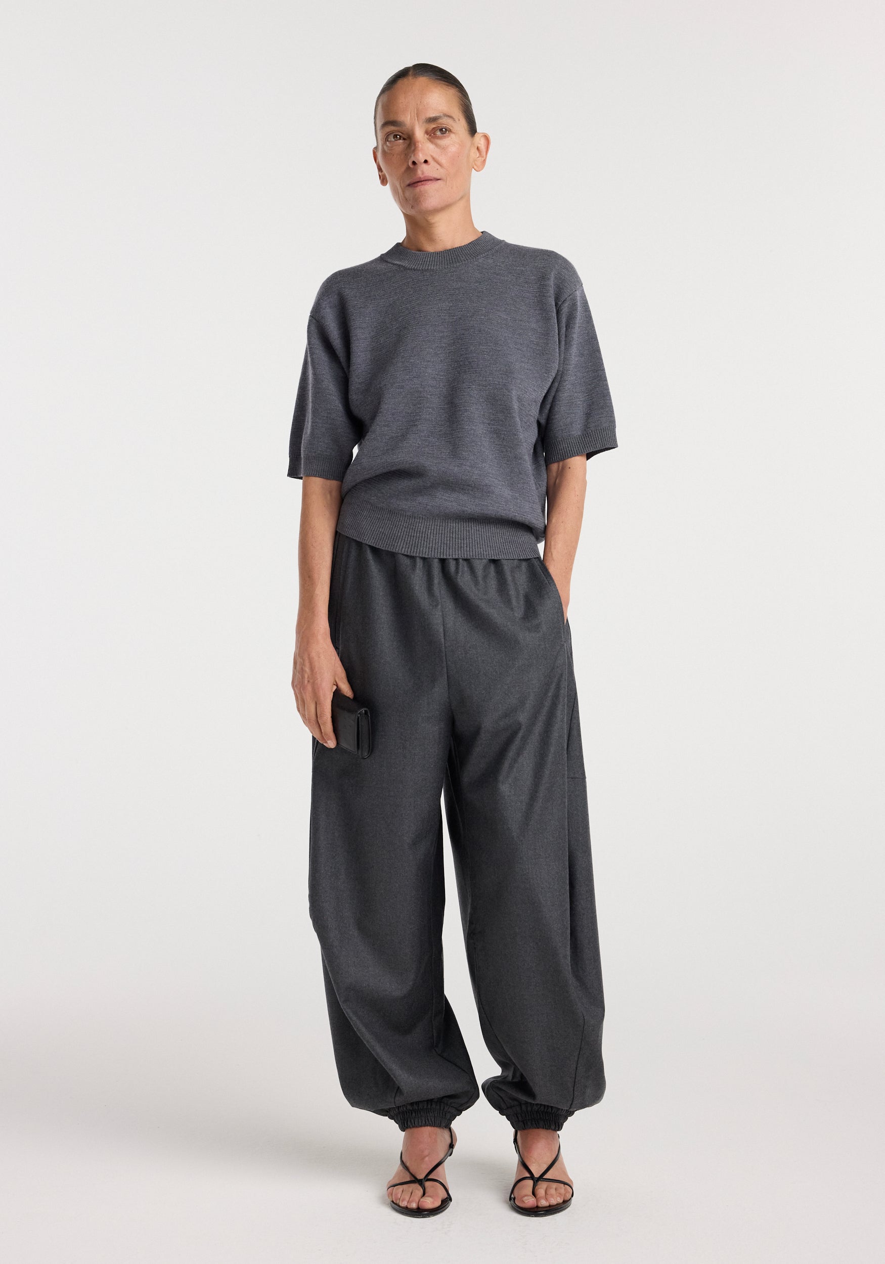 Woman wearing a dark grey melange compact knitted top with short sleeves, paired with loose dark grey pants and black open-toe sandals, standing against a plain light background.