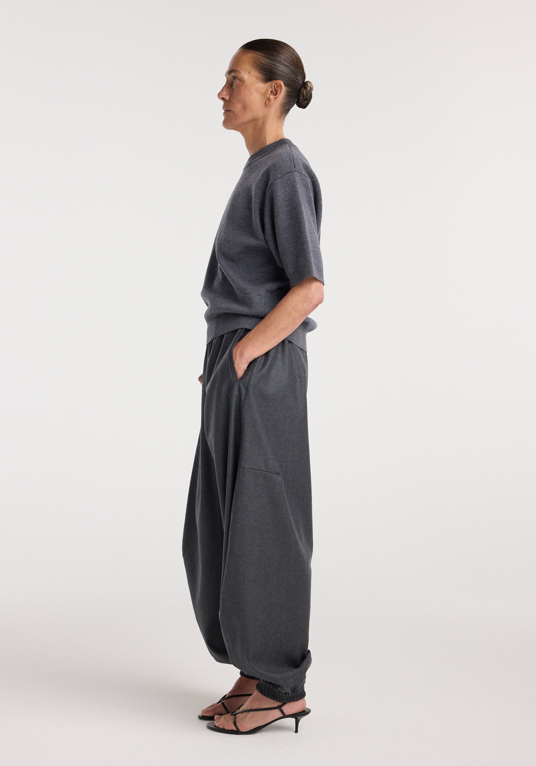 Side view of a woman wearing a dark grey melange compact knitted top with short sleeves, paired with loose dark grey pants and black strappy heels against a plain light background.