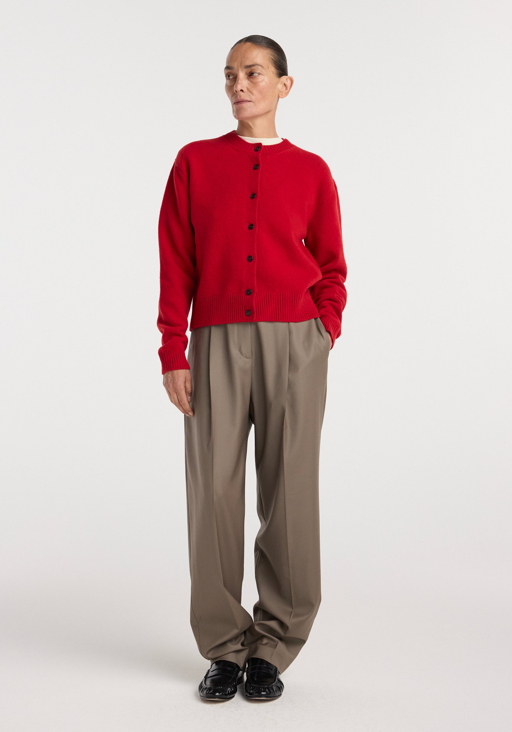 Woman wearing a red button-up Classic cardigan | red paired with loose beige trousers and black shoes, standing against a plain light background.