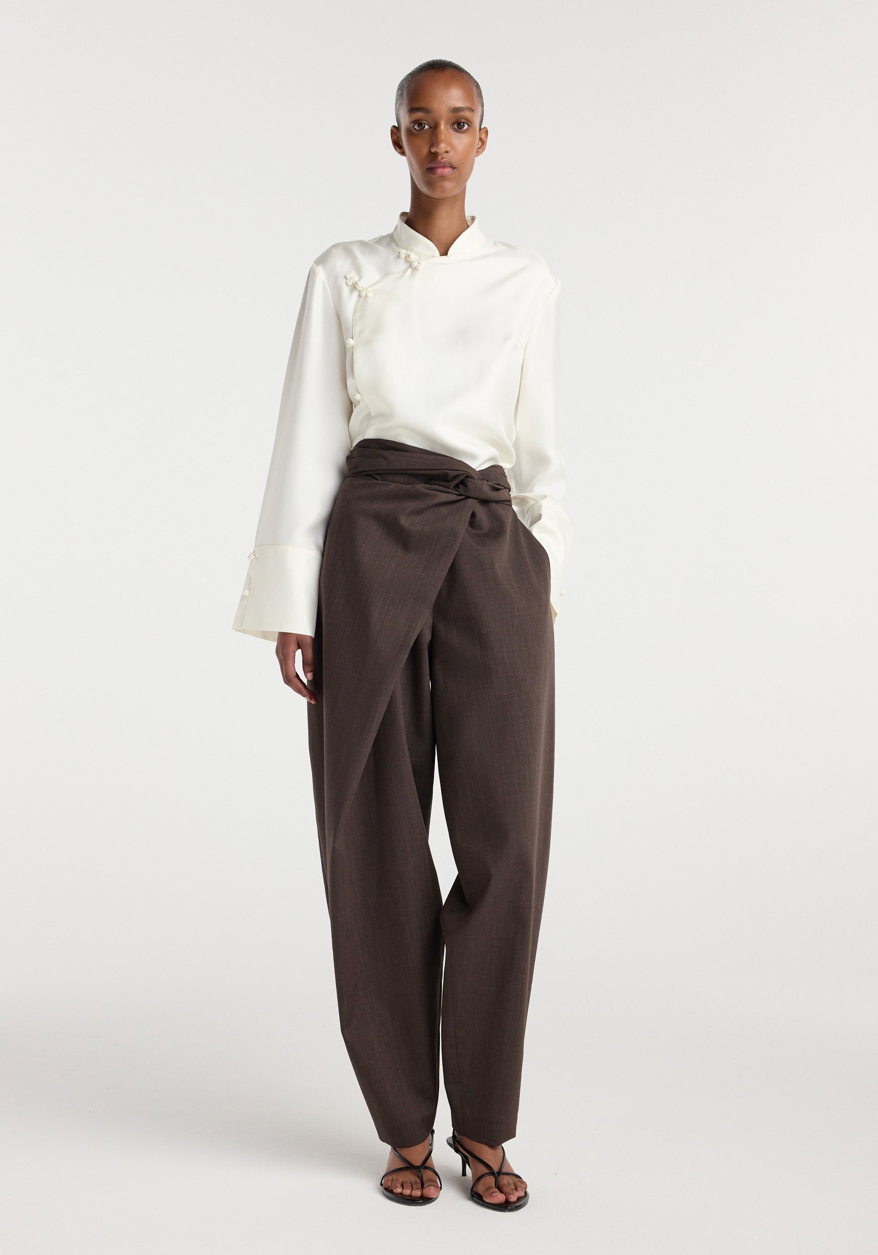 Model wearing Sarong knot trousers | dark brown paired with a white long-sleeve blouse and black strappy sandals, standing against a plain light background.