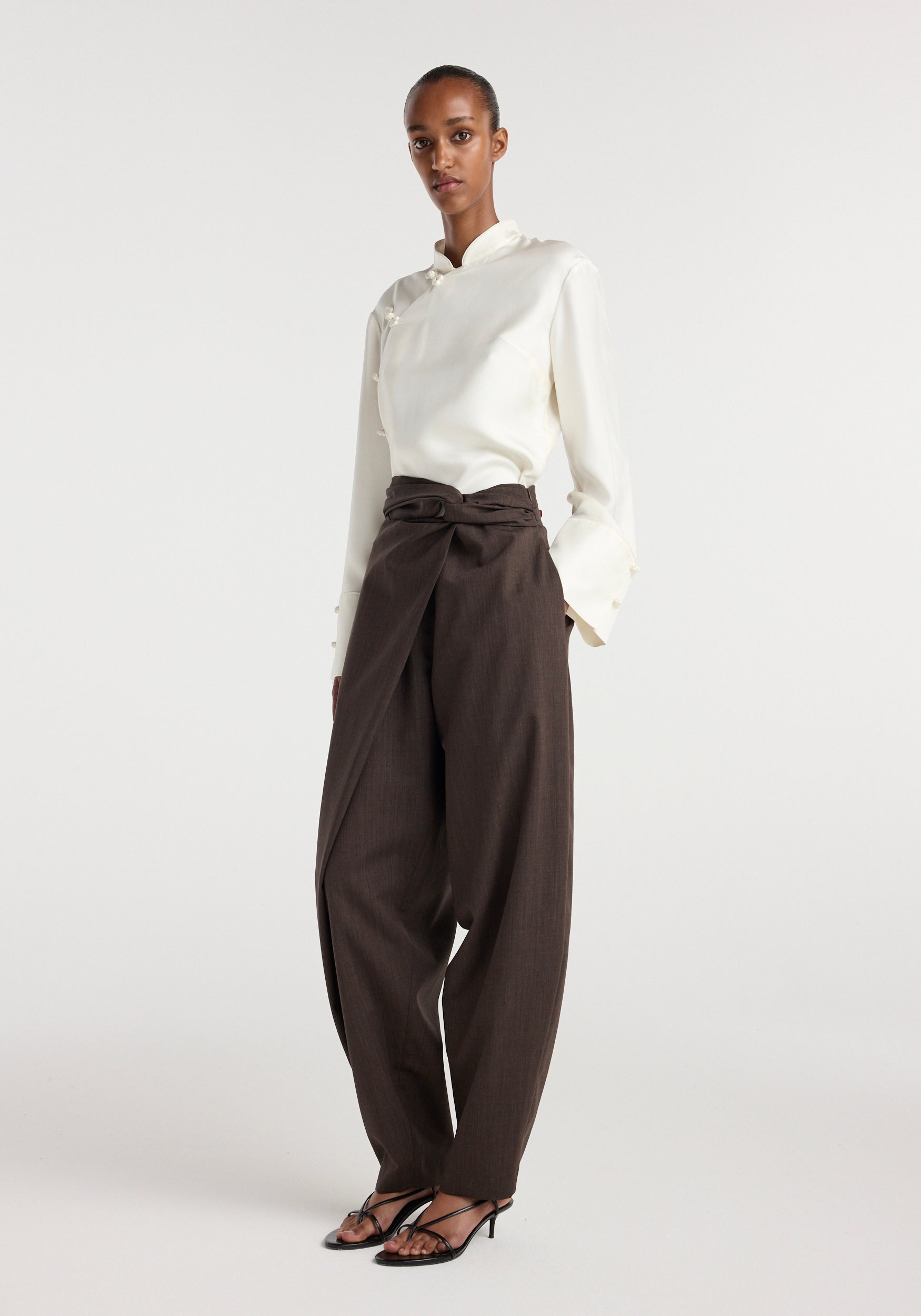 Model wearing Sarong knot trousers | dark brown with a high waist and wide legs, styled with a white long-sleeve top and black strappy heels against a plain background.