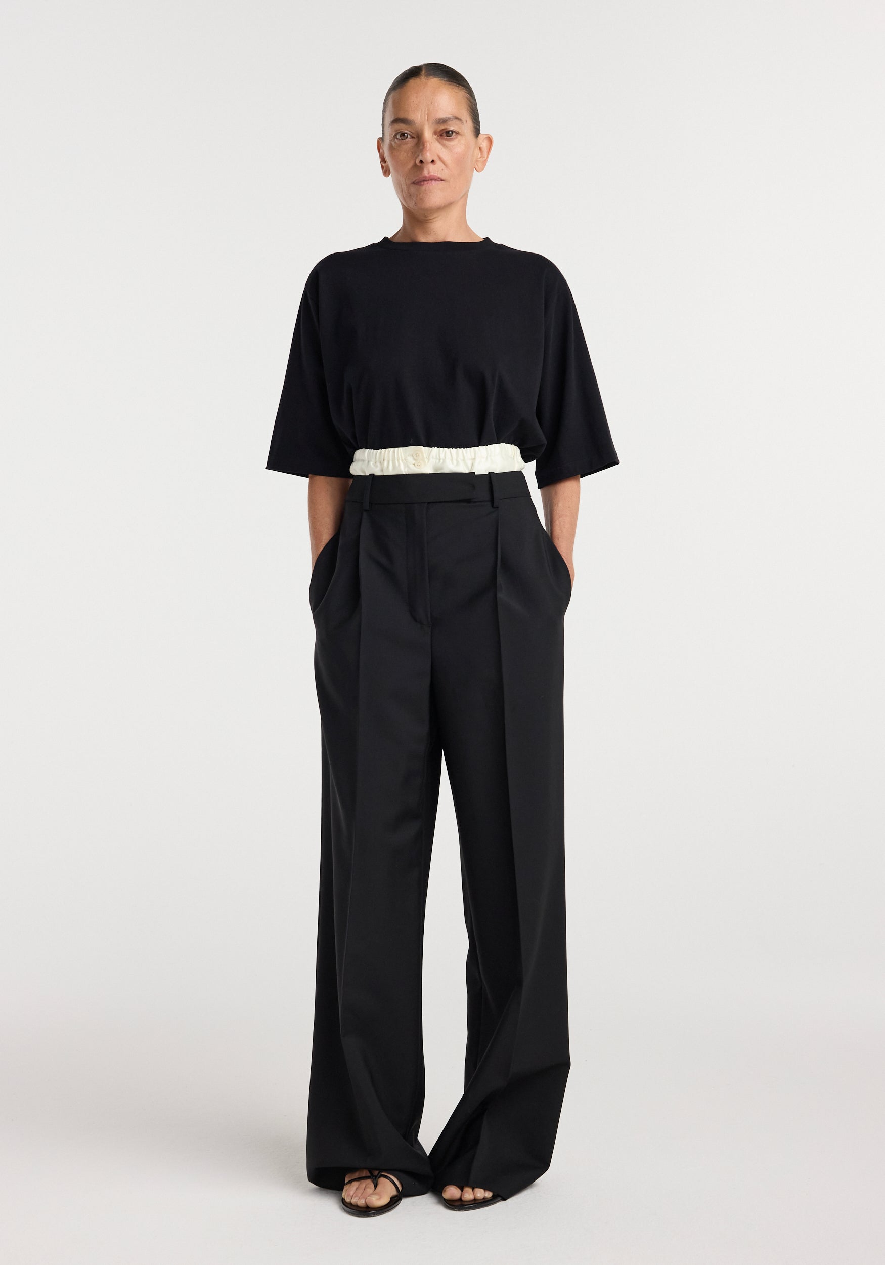 Woman wearing black Boxer detailed trousers | black with a high waist, wide-leg cut, side pockets, and a white elastic belt, paired with a loose black top and black sandals.