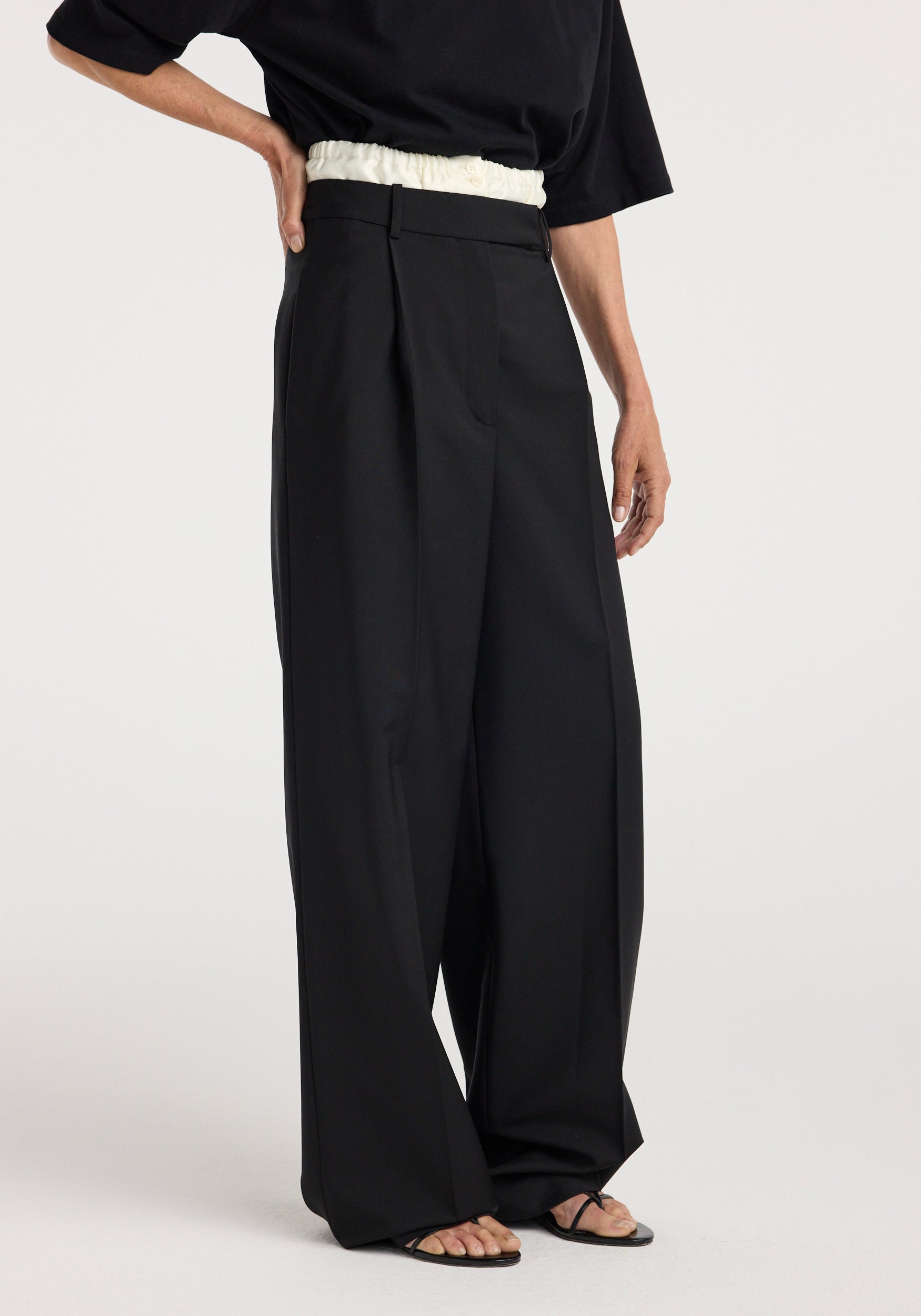 Side view of a person wearing black wide-leg Boxer detailed trousers | black with a high-waist elastic white waistband, paired with a loose black top and black open-toe sandals.