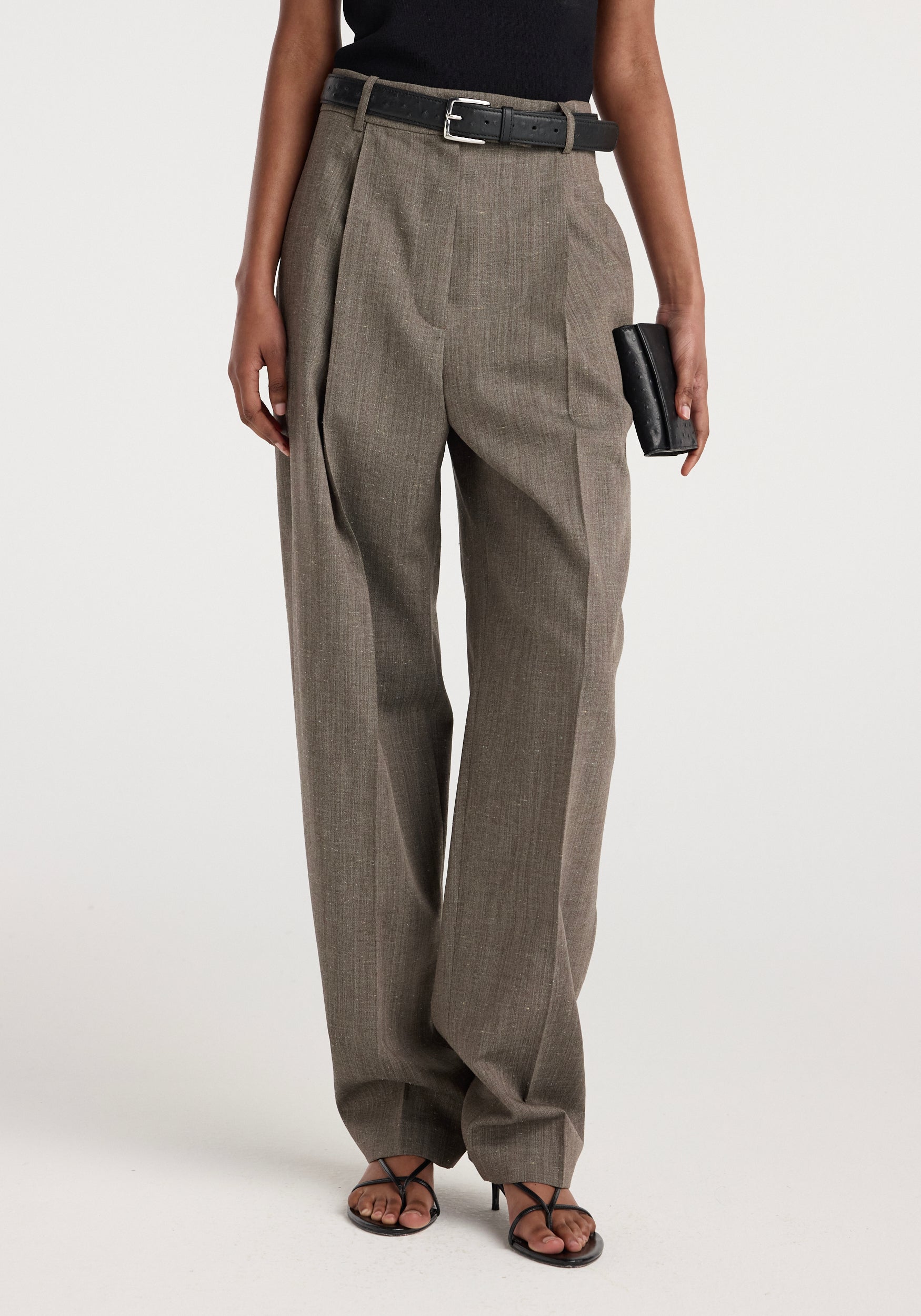 Front view of a person wearing Daria - Single pleat tapered trousers | earth slub in a muted brown tone, paired with a black belt, black sleeveless top, black clutch, and black open-toe heels.