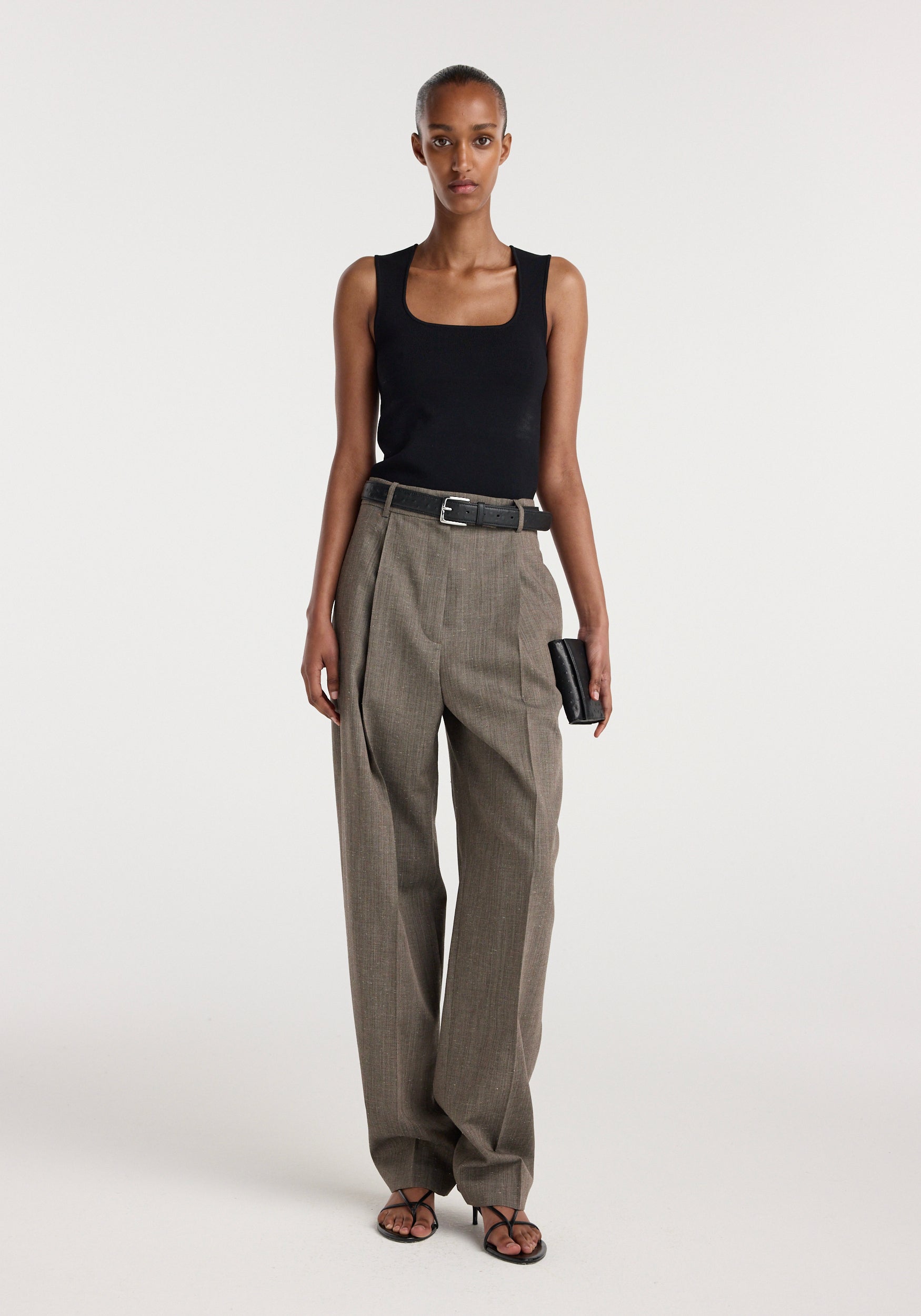 Woman wearing a black sleeveless top paired with Daria - Single pleat tapered trousers | earth slub, featuring a belted waist and straight-leg fit, standing against a plain background.