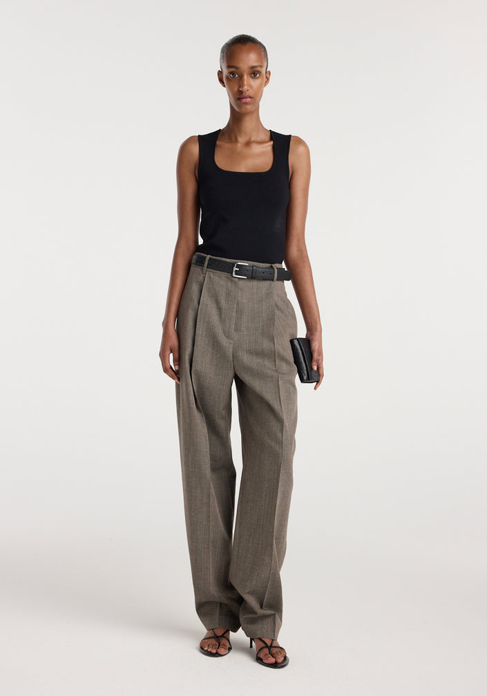 Woman wearing a black sleeveless top paired with Daria - Single pleat tapered trousers | earth slub, featuring a belted waist and straight-leg fit, standing against a plain background.