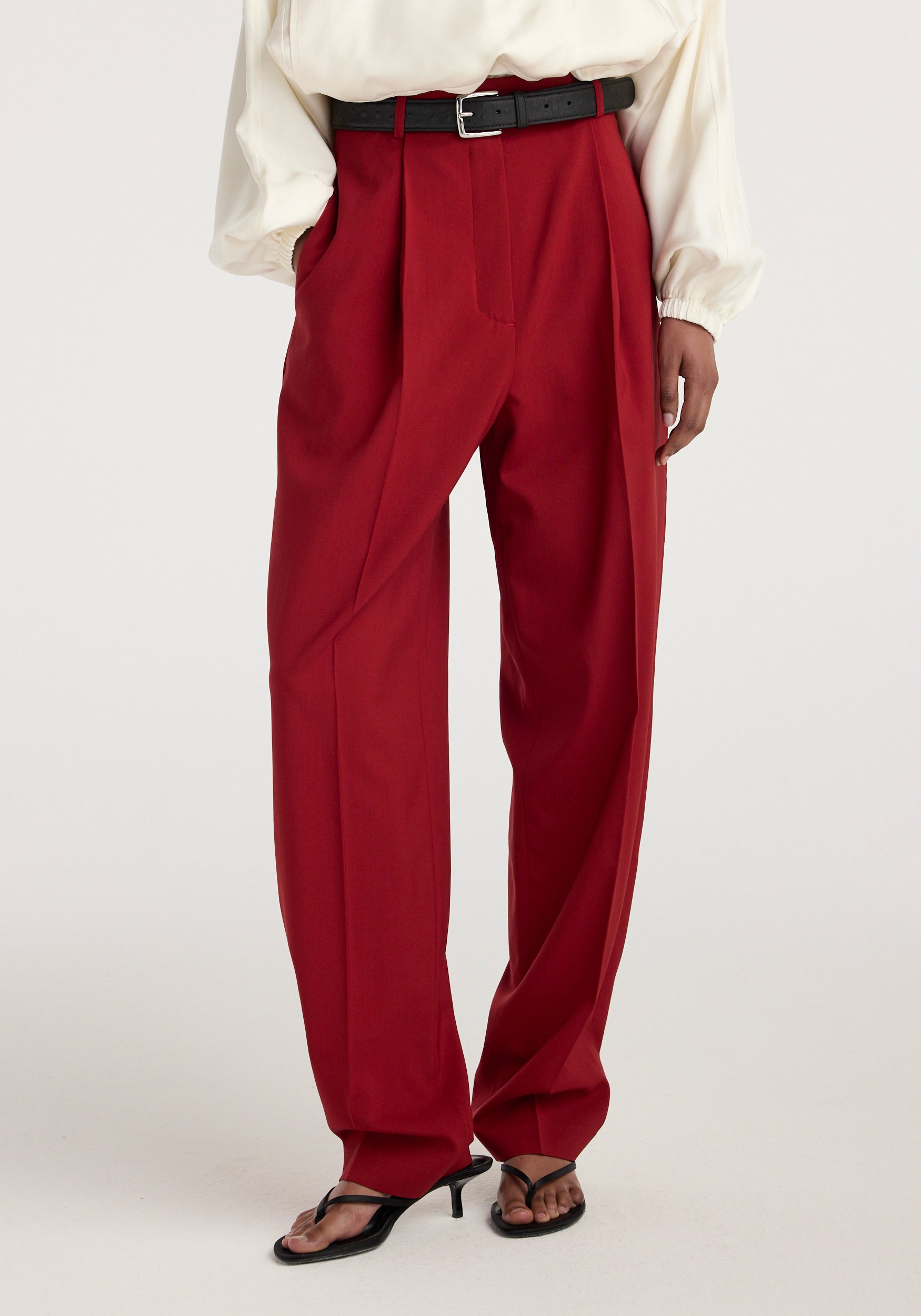 Front view of a person wearing Daria - Single pleat tapered trousers | carmine in deep red, styled with a black belt, cream blouse, and black heeled sandals.