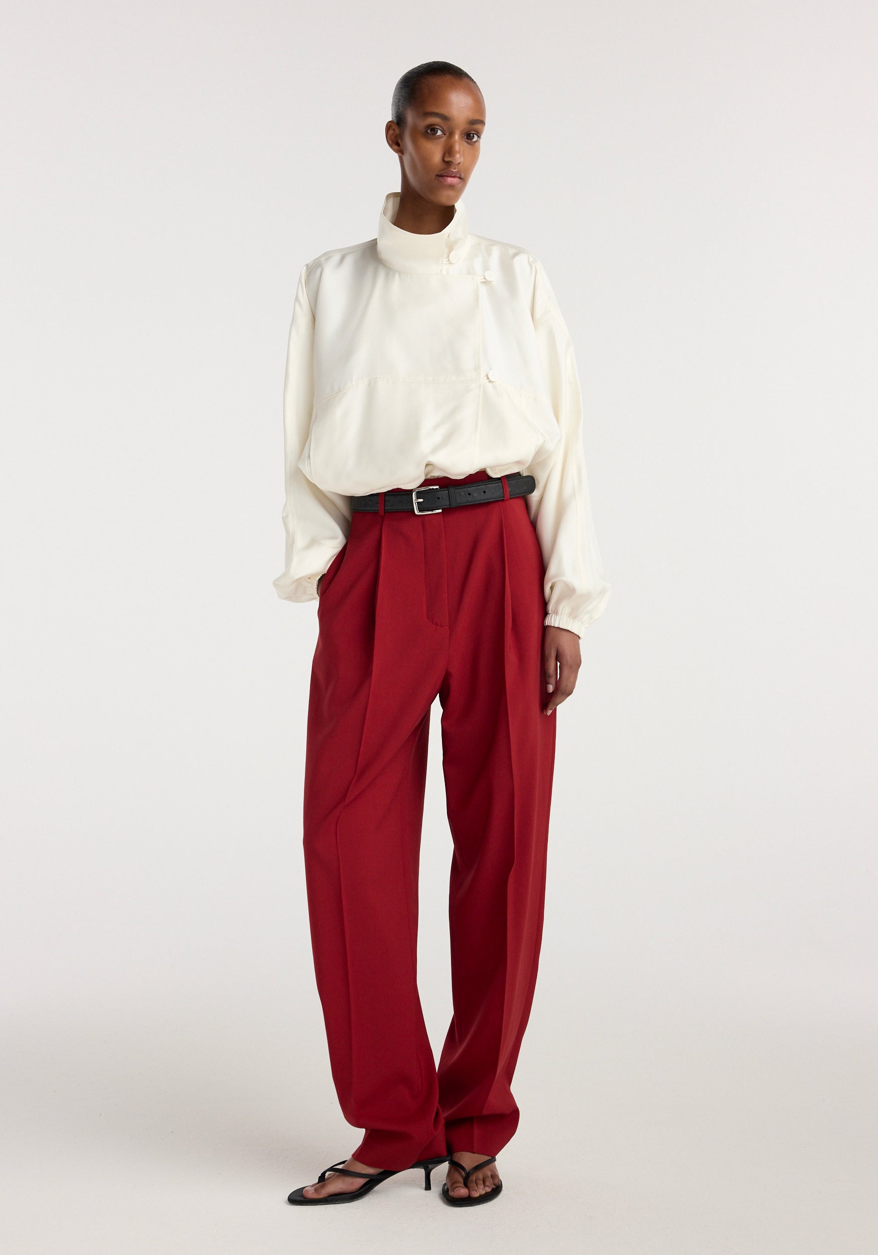 Model wearing Daria - Single pleat tapered trousers | carmine in a rich red shade, styled with a high-waisted fit, pleated front, and paired with a white long-sleeve blouse and black belt.