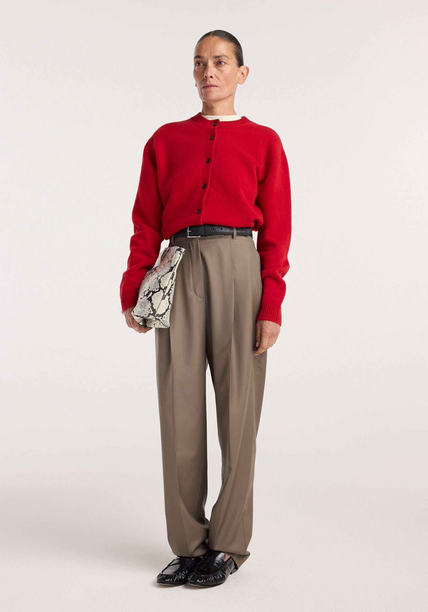 A model wearing Daria - Single pleat tapered trousers | desert brown paired with a red button-up sweater, black belt, black shoes, and holding a snakeskin-patterned clutch bag.