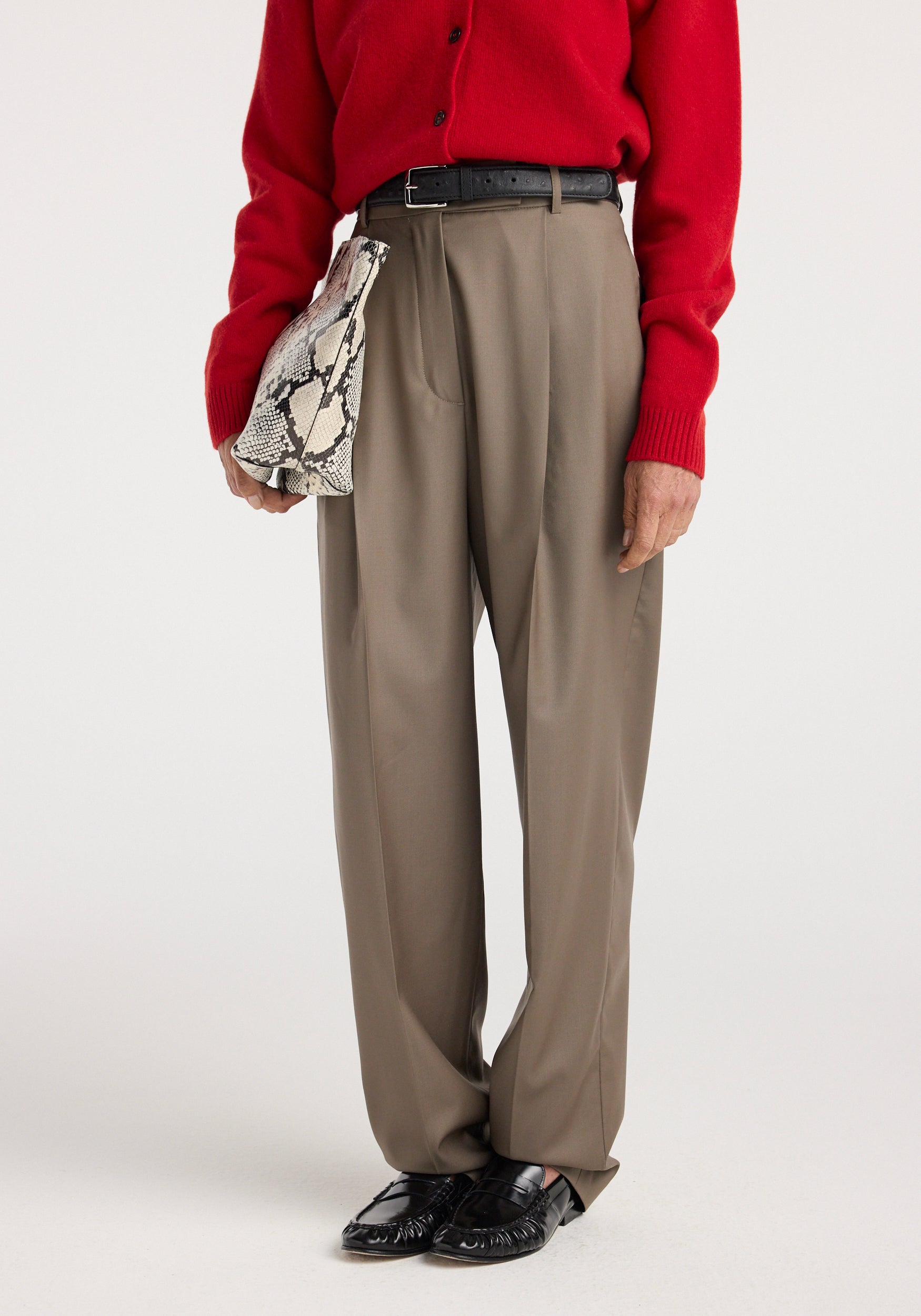 A person wearing Daria - Single pleat tapered trousers | desert brown paired with a red sweater, black belt, black loafers, and holding a snakeskin patterned handbag.