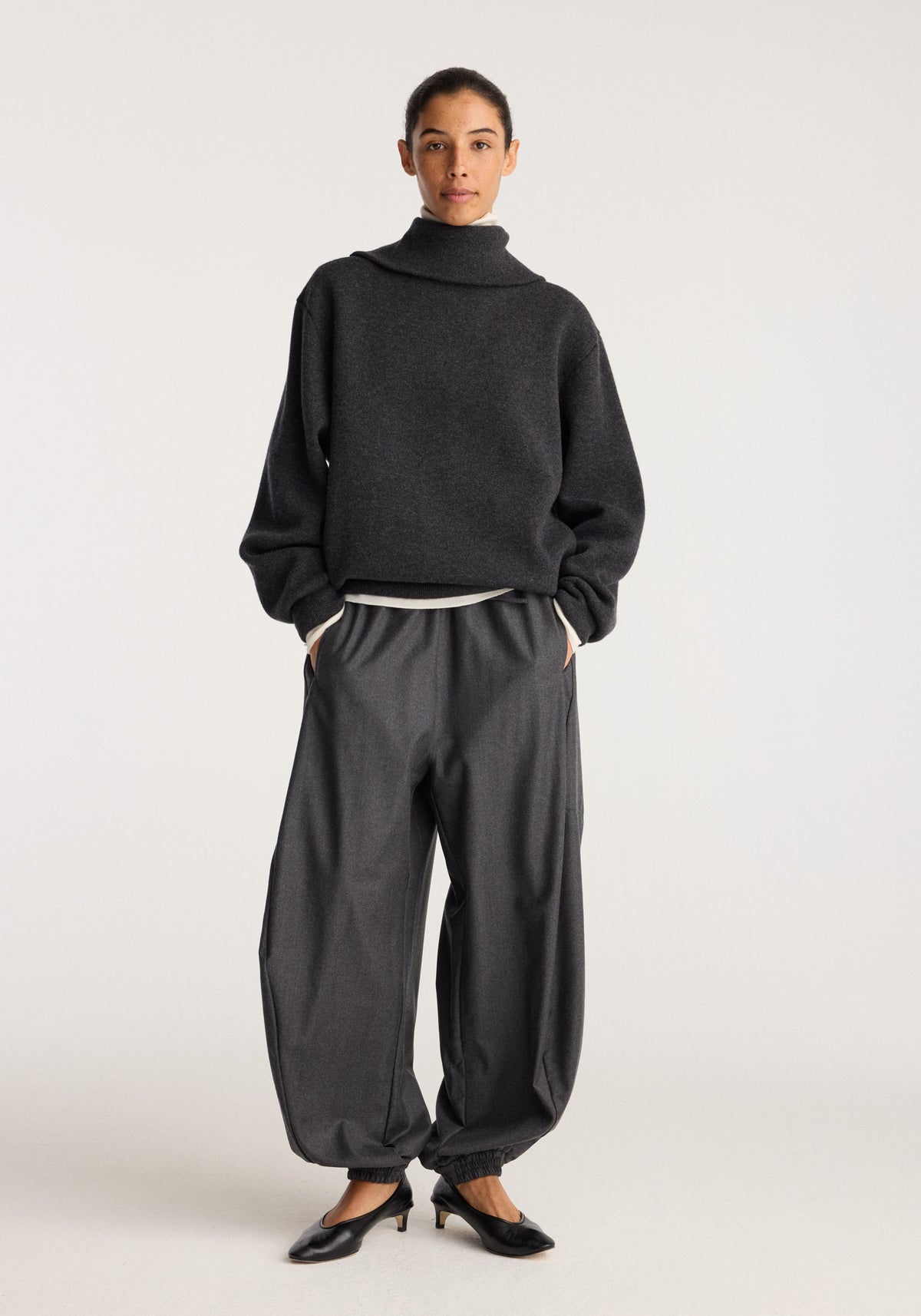 Balloon track pants | dark grey melange worn by a model with a loose fit, elastic waistband, tapered legs, and ankle cuffs styled with a dark sweater and loafers