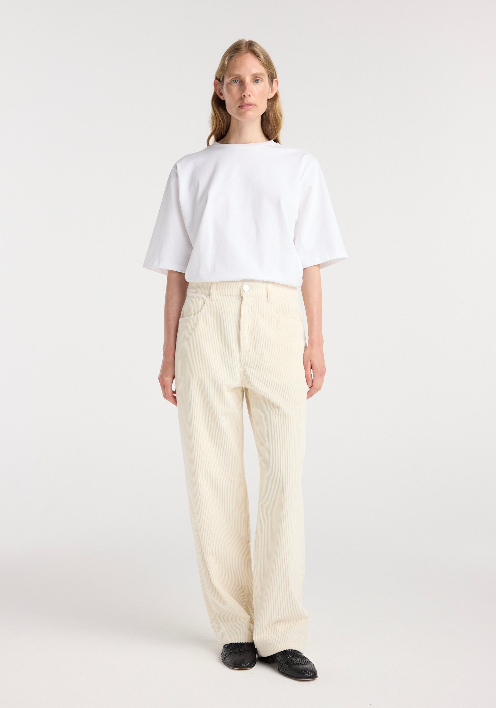 Woman wearing winter white Bruun - Signature baggy corduroy pants paired with a plain white oversized short-sleeve t-shirt and black shoes against a white background.