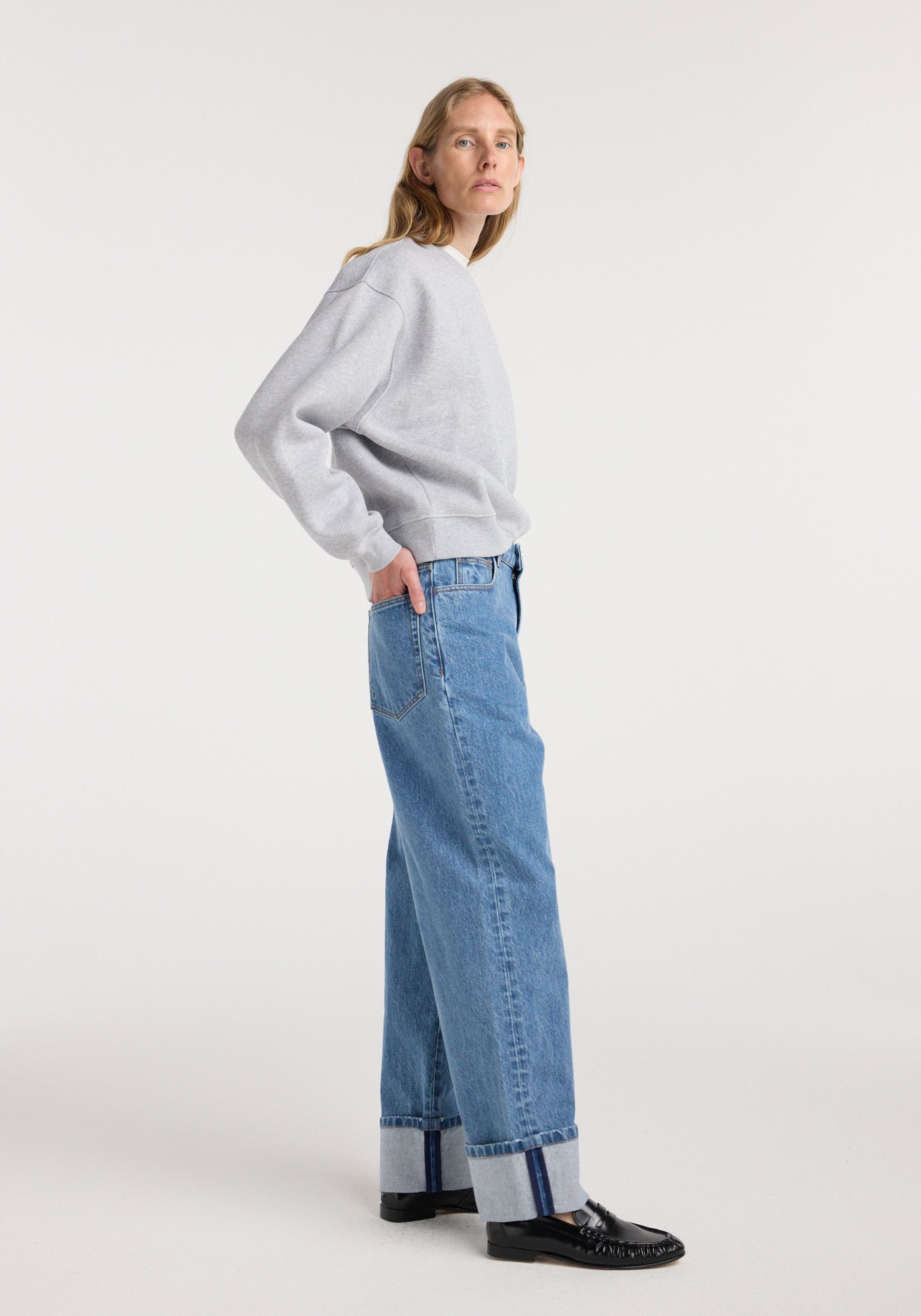 Woman wearing Rixt - Turn-up baggy denim | heritage blue with a grey sweatshirt and black shoes, standing in a side profile against a plain white background.