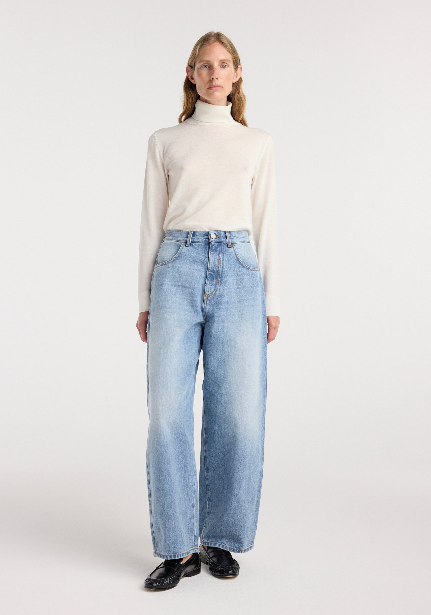 Woman wearing Berber - Barrel leg denim | classic blue, styled with a cream turtleneck sweater and black shoes in a minimalist studio setting.