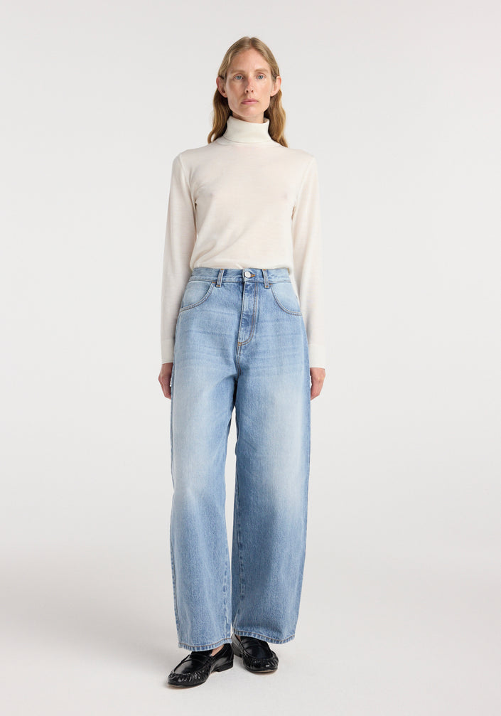 Woman wearing Berber - Barrel leg denim | classic blue, styled with a cream turtleneck sweater and black shoes in a minimalist studio setting.